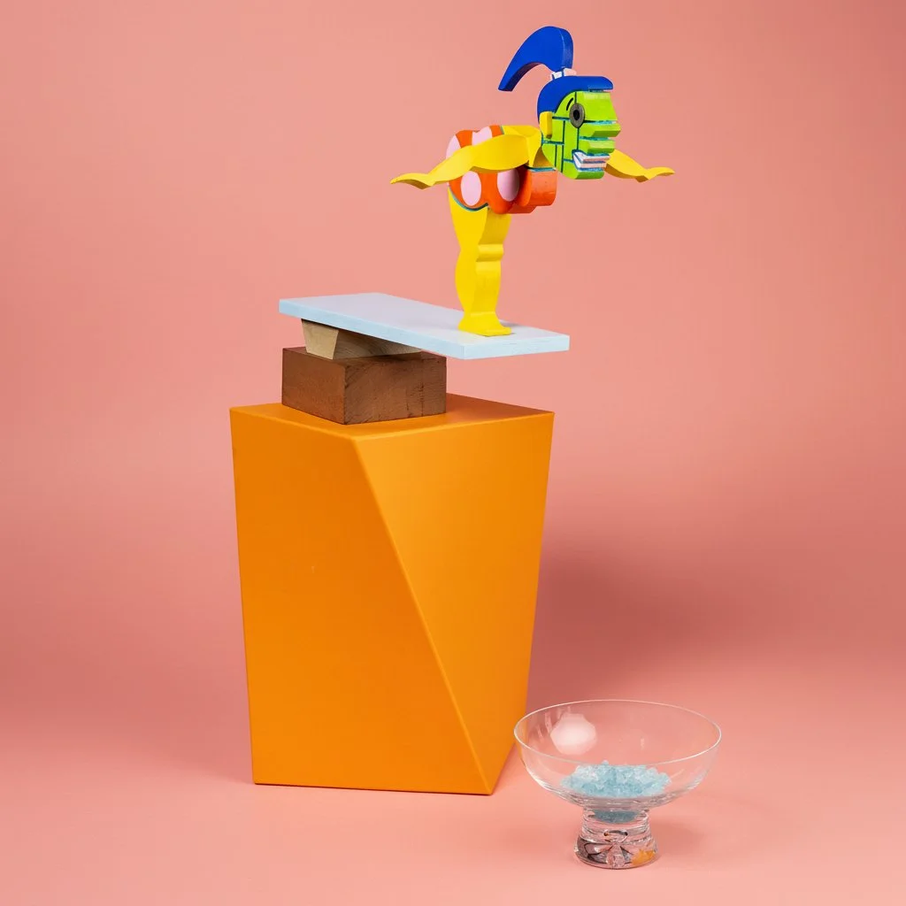 Colorful abstract sculpture on a yellow and white platform against a pink background, with a glass bowl filled with ice and a white object nearby.