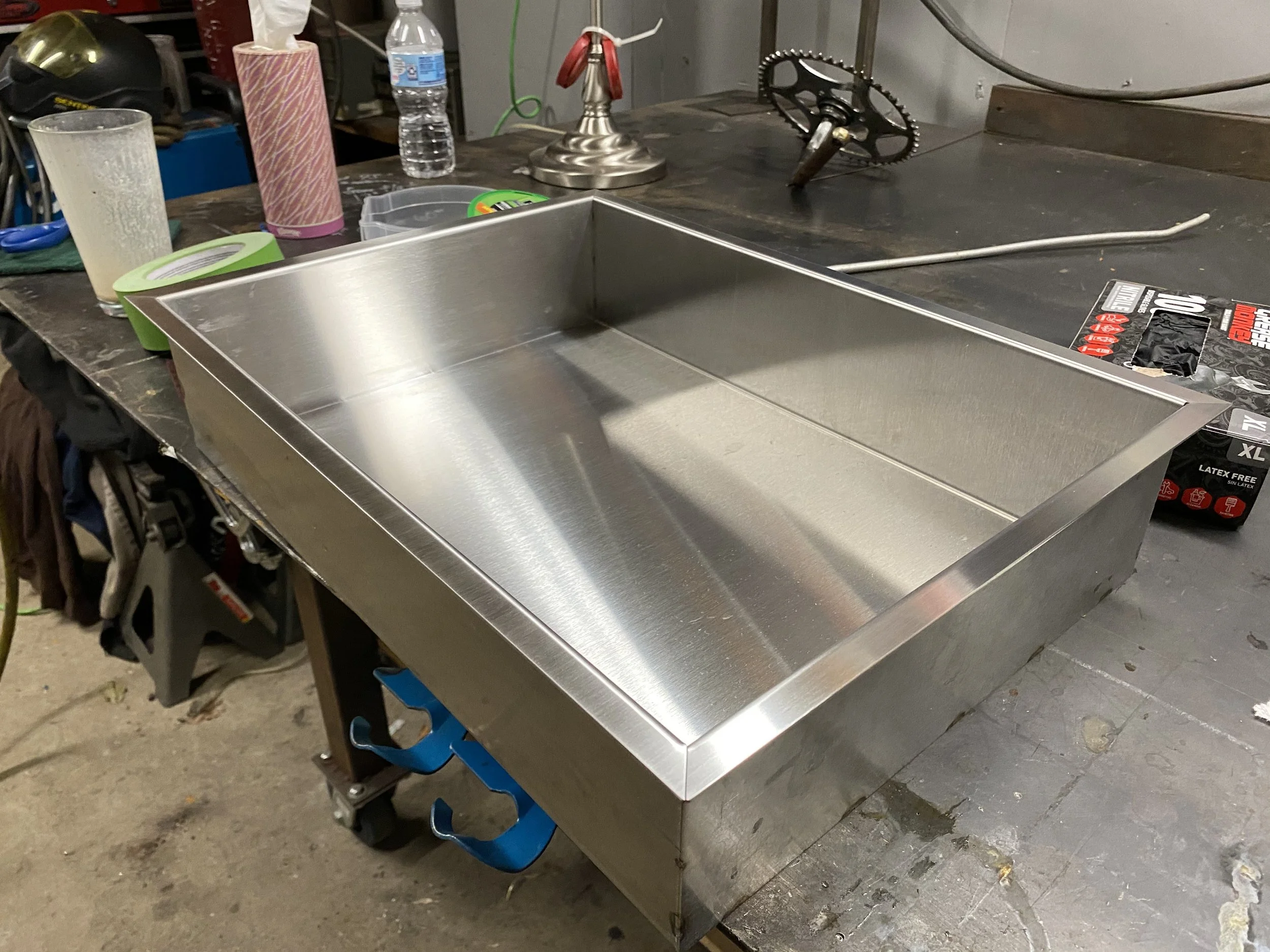 A stainless steel rectangular fire pit or cooking station on a workbench in a workshop, surrounded by tools, tape, a water bottle, and various workshop items.