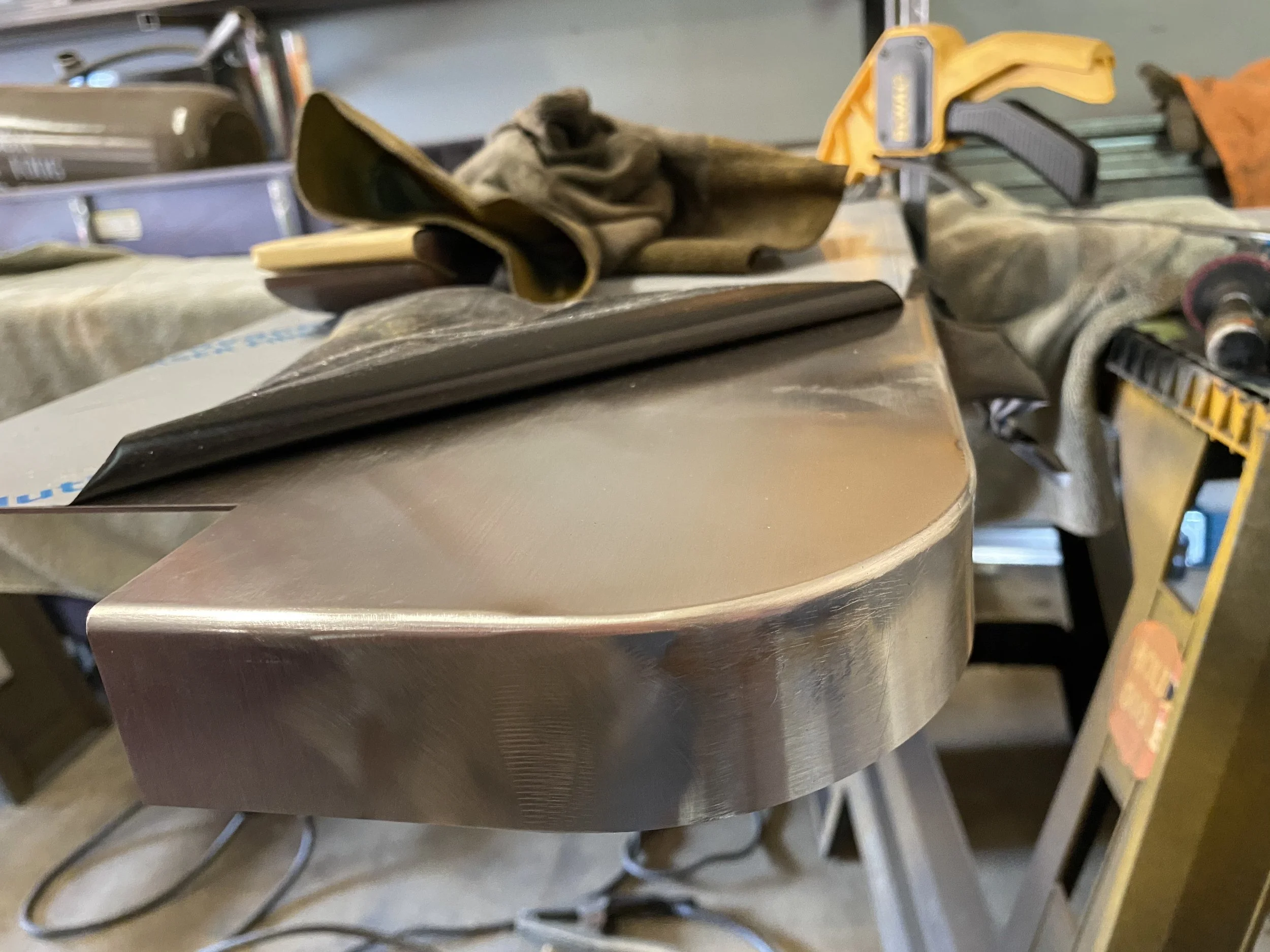 A close-up view of a metal workbench with various tools and materials, including a clamp, gloves, and a black cloth, in a workshop setting.