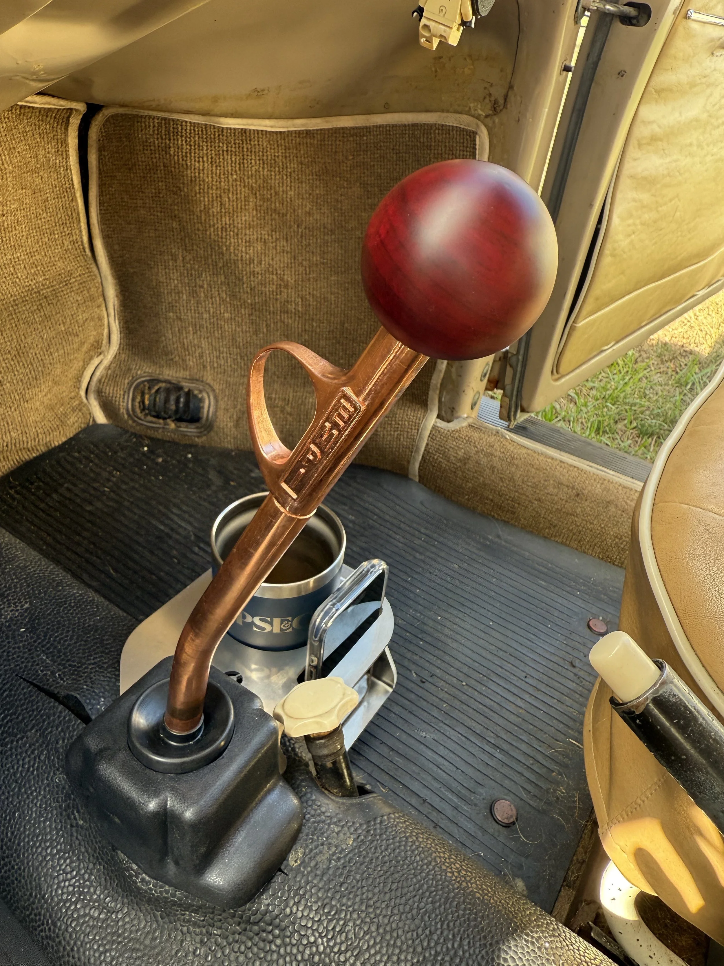 Gear shift lever with a wooden ball knob inside an older vehicle with tan interior and black rubber mat.