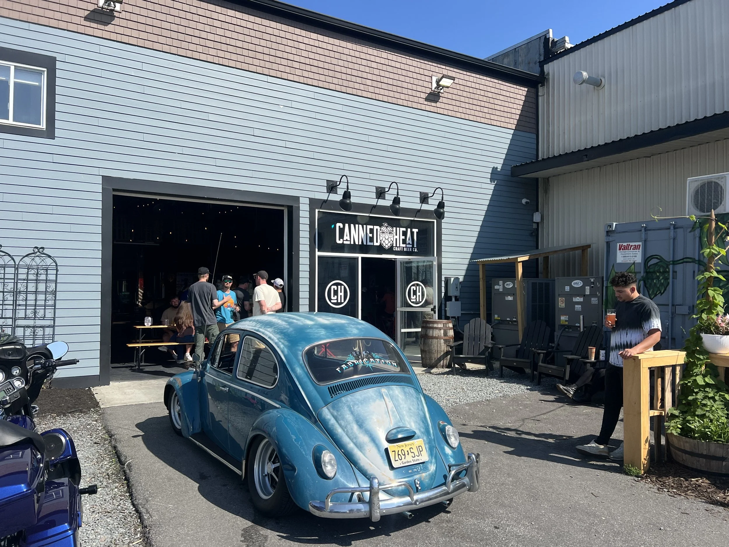 Outside of Canned Heat craft beer bar with a vintage blue Volkswagen Beetle parked in front. People are gathered inside and outside, some holding drinks.