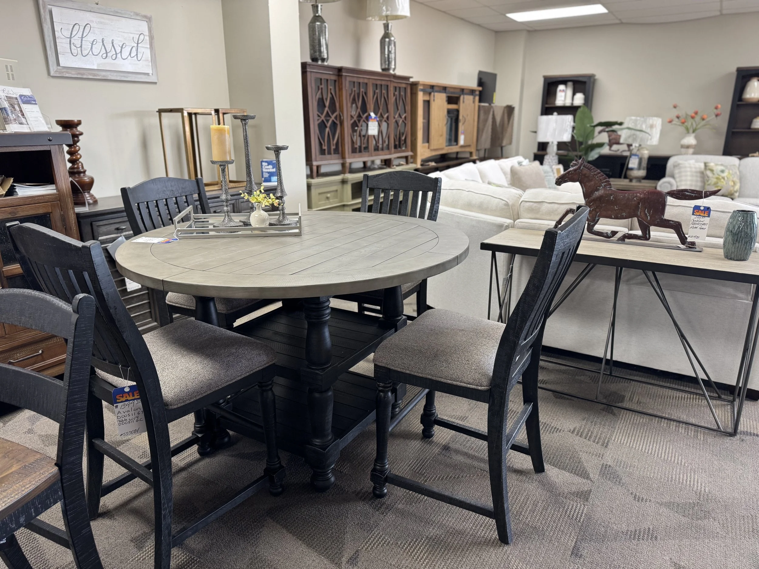 Furniture display in a store, including a round dining table with five black chairs, a wooden console table with a horse figurine, and various cabinets and decorative items in the background.