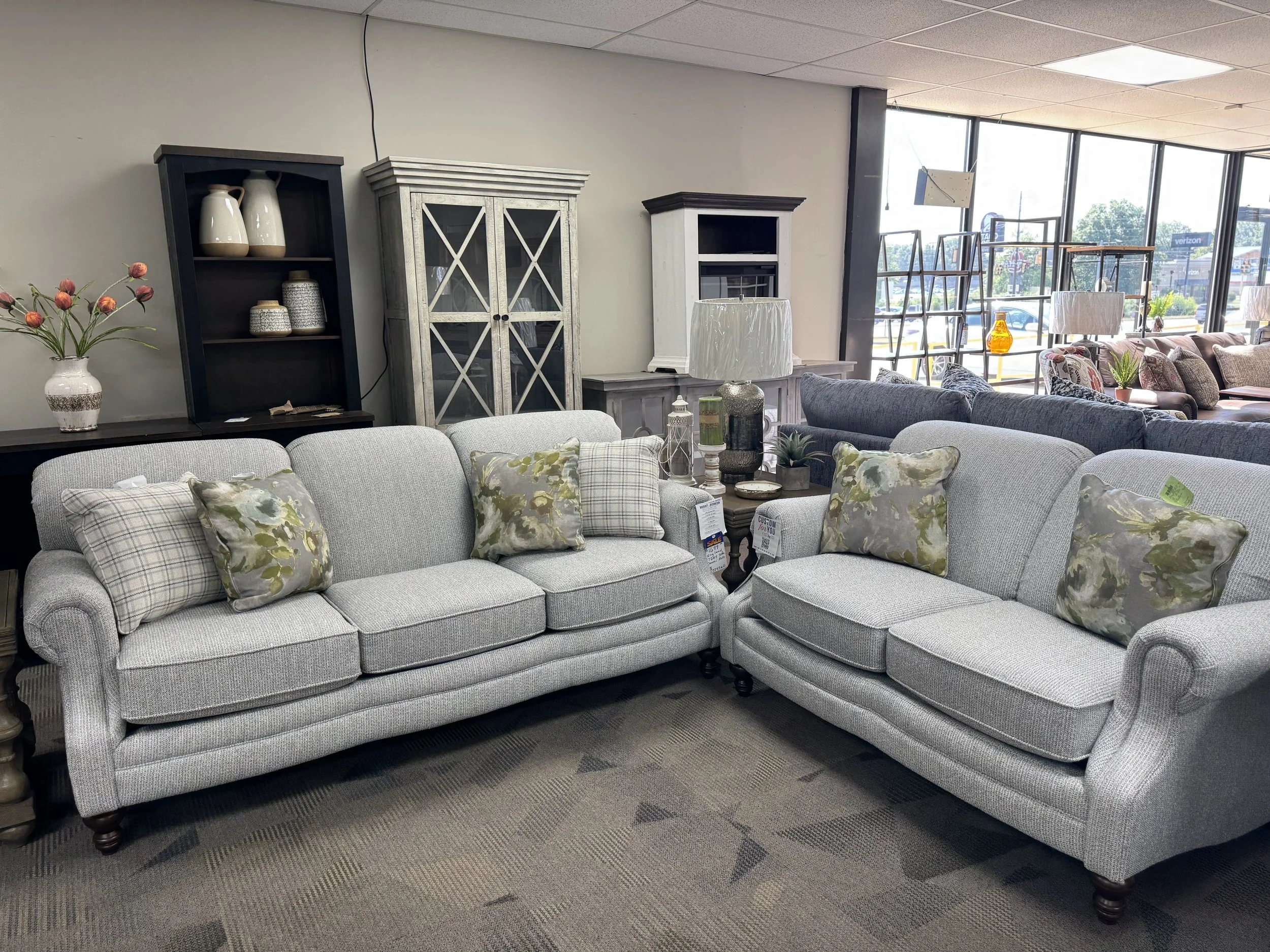 A furniture store with two light gray upholstered sofas with patterned throw pillows, a black shelf with decorative items, and large windows letting in natural light.
