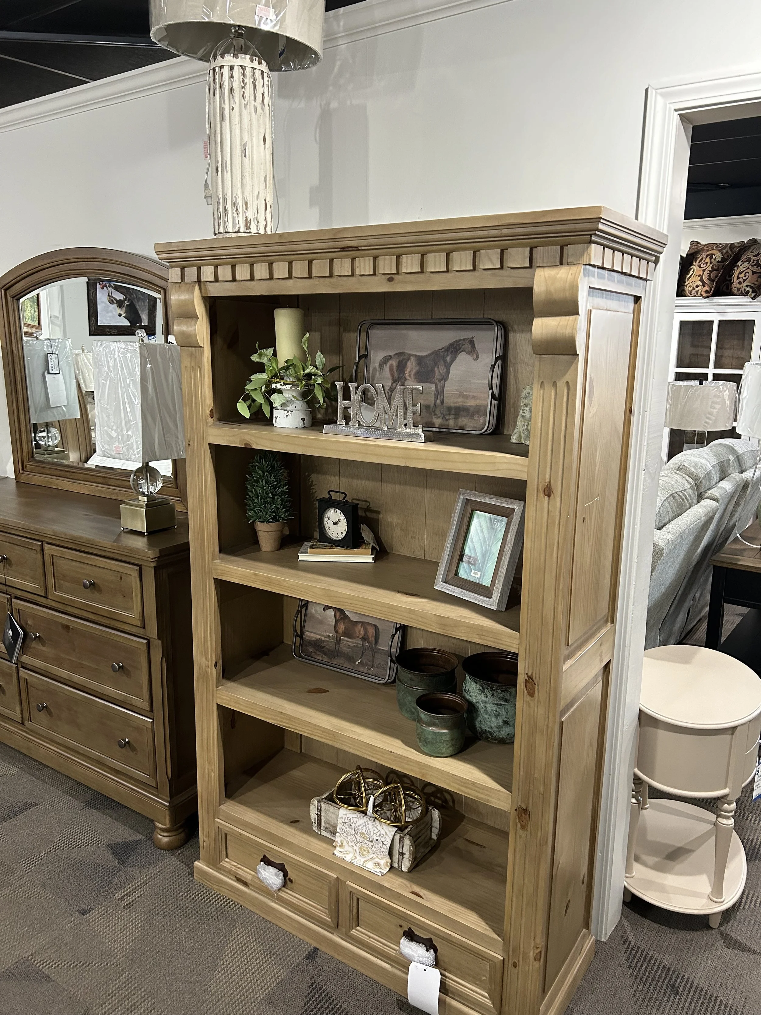 Wooden bookshelf with decorative items, including a metal "HOME" sign, framed picture of a horse, small potted plant, clock, and ceramic pots, in a furniture store.