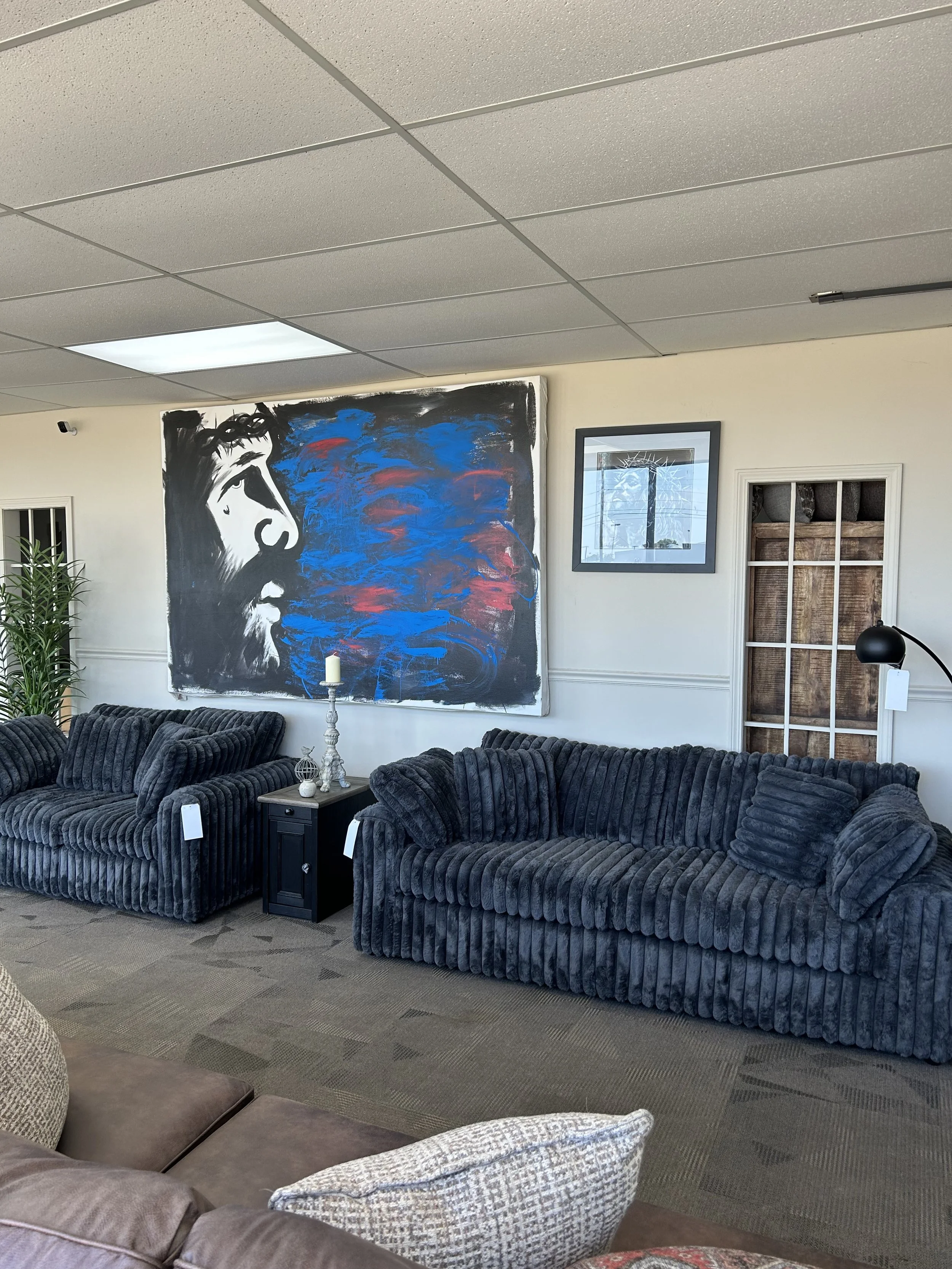 A living room with dark gray velvet sofas, a black side table with decorative items, a large abstract painting of a face with blue and red elements, framed artwork, a window, a lamp, and a door with a wooden frame.