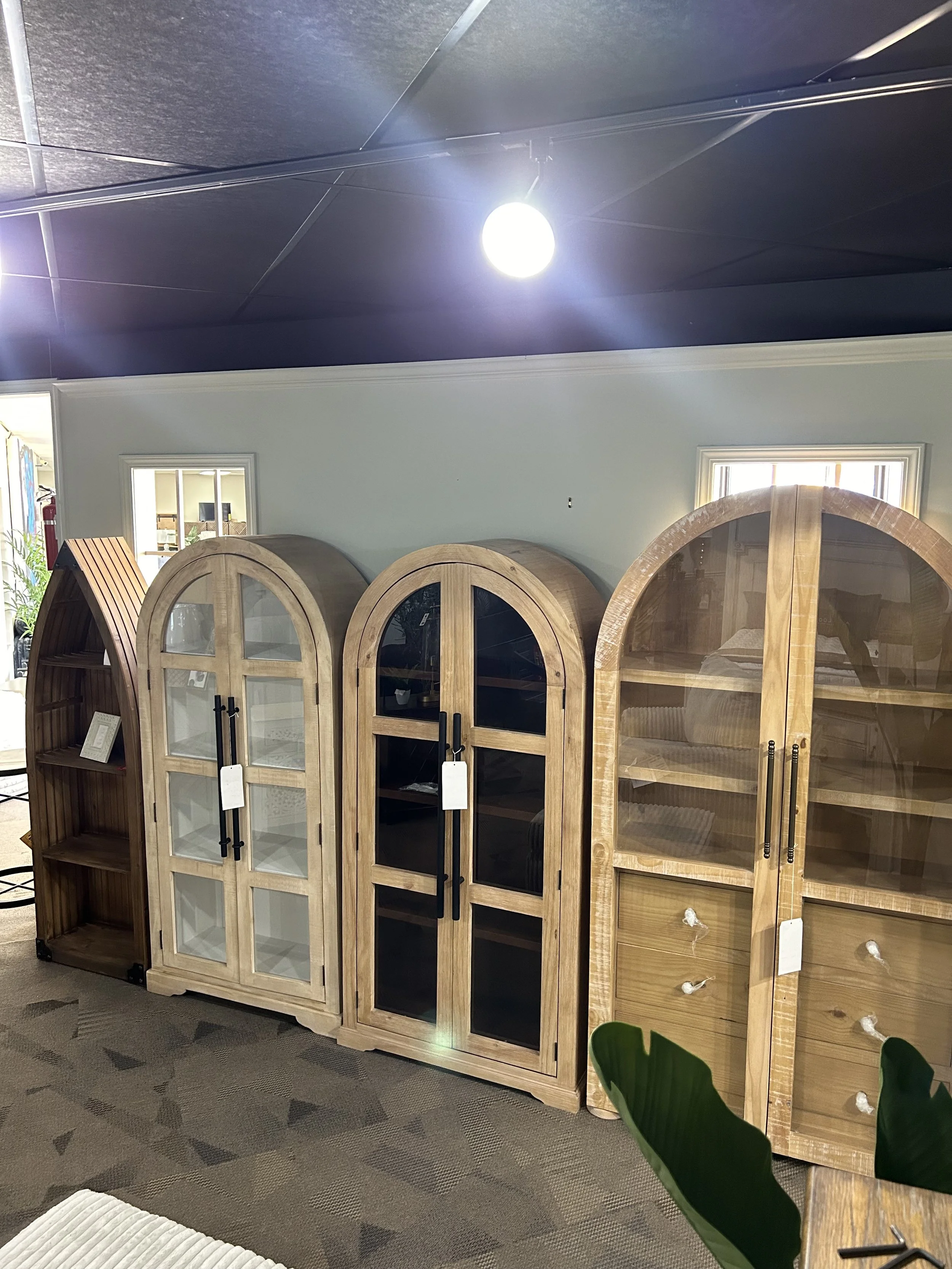 Three wooden display cabinets with glass doors and black handles, positioned side by side in a store, with a small window behind them. The cabinet on the right has glass knobs on its drawers, and there are some green leaves visible in the bottom righ