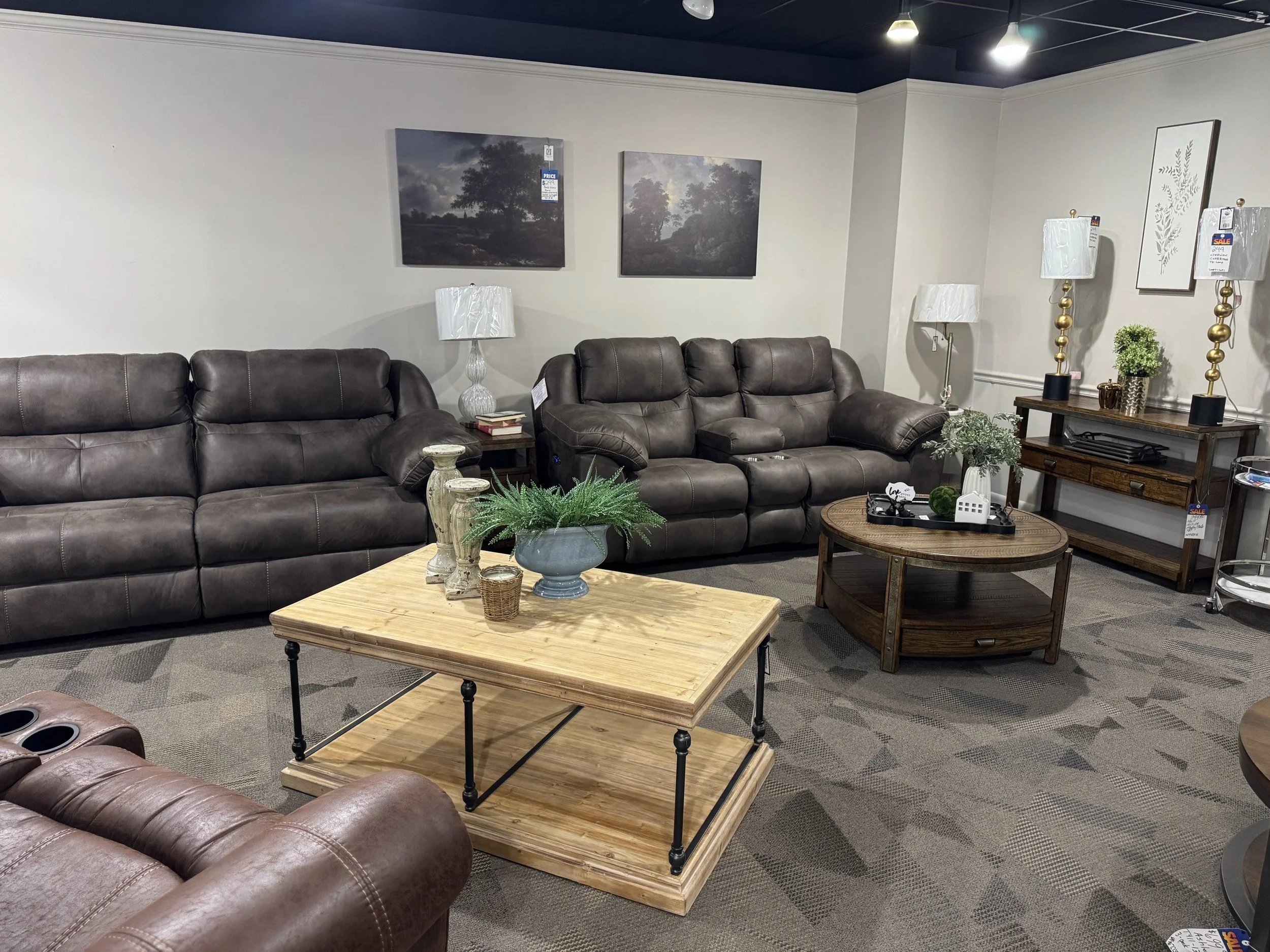 A furniture display in a home furniture store includes three dark brown leather sofas, a light wood coffee table with decorative green plants and candles, and a wooden console table with matching table lamps, framed artwork, and decorative plants on