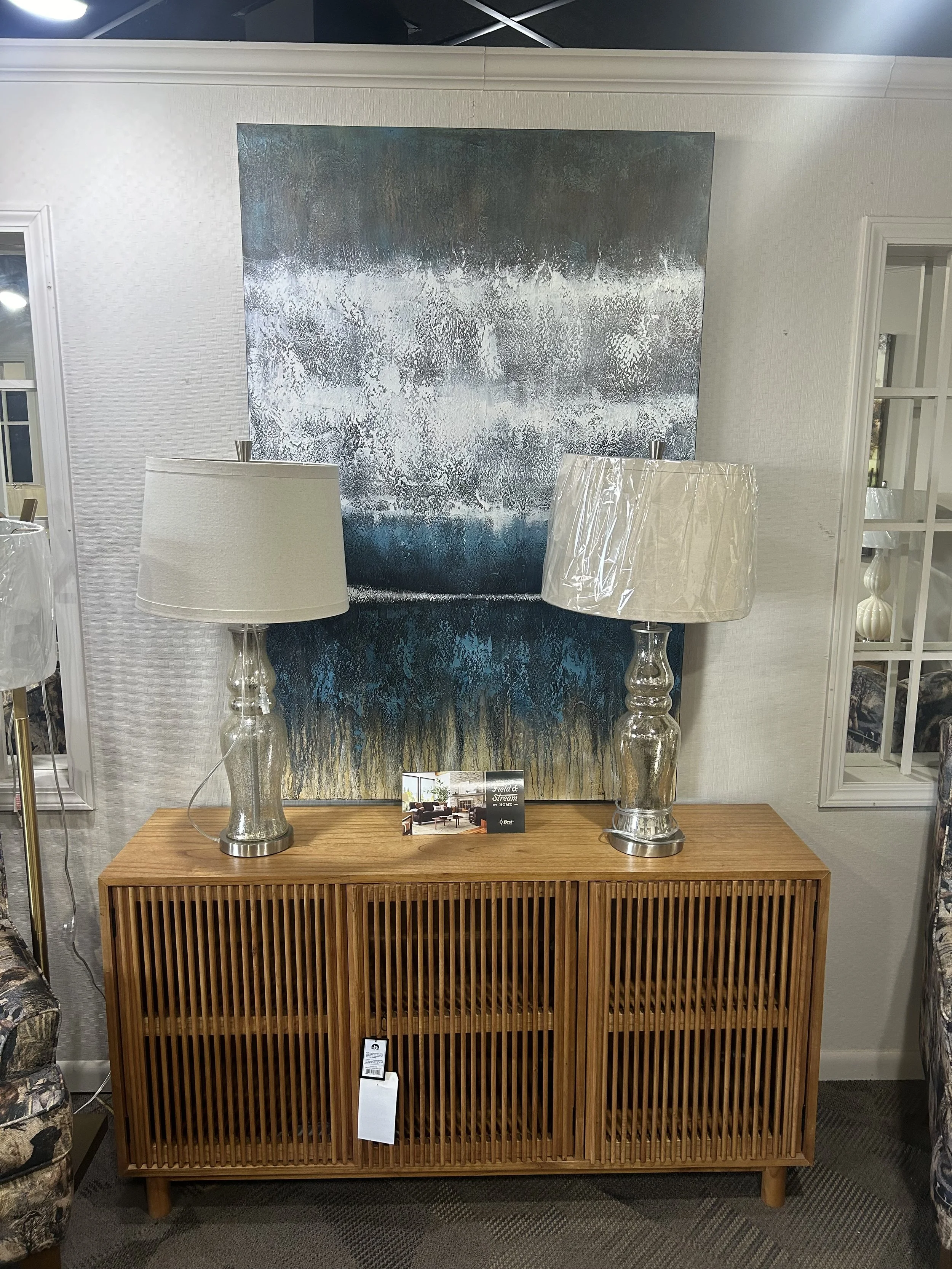 A wooden sideboard with slatted doors, two large glass table lamps with white shades, and a large abstract painting with blue, gray, brown, and white textures hanging on the wall behind it.