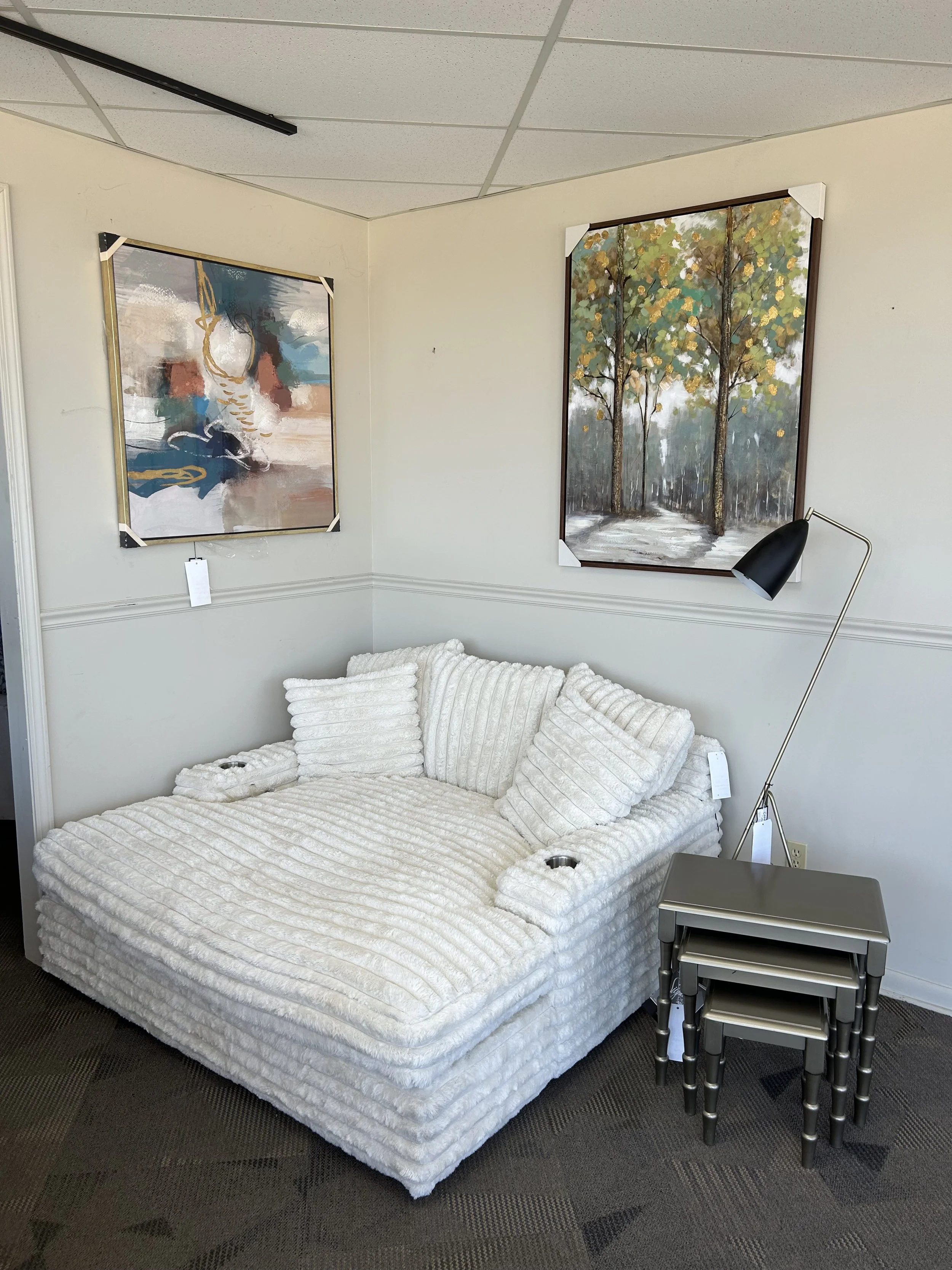 A living room corner with a white, corduroy-textured sectional sofa with multiple pillows, a small nesting table set, a modern black floor lamp, and two paintings on the wall depicting abstract art and a landscape with trees.