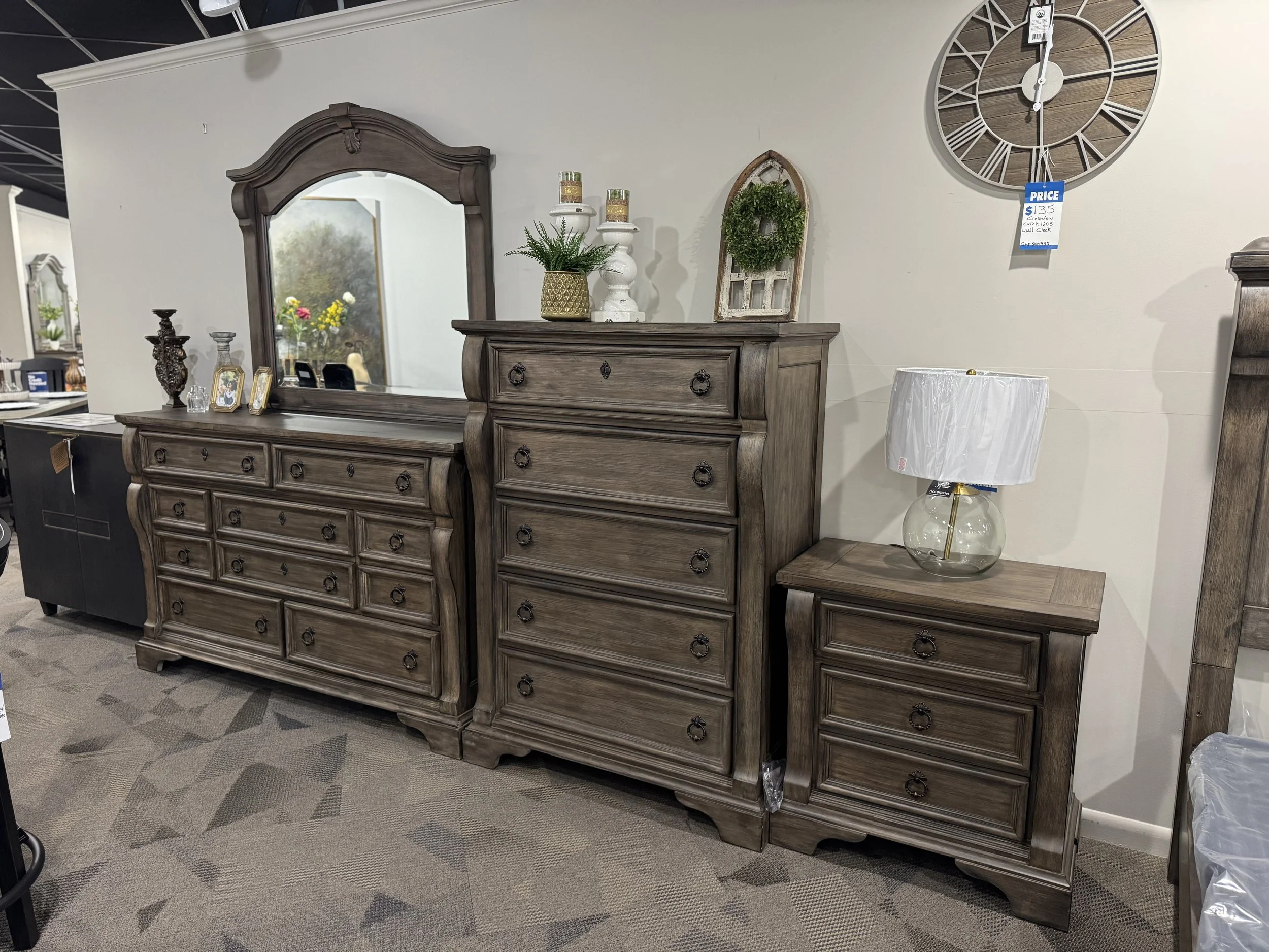 Set of three brown wooden dressers with drawers, on a carpeted floor, accompanied by a glass table lamp, decorative items, and a large wall clock.