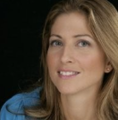 A woman with long, light brown hair and a slight smile, wearing a blue top against a dark background.