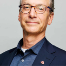 Portrait of a middle-aged man with glasses, wearing a navy blazer and a blue shirt, smiling slightly against a light gray background.
