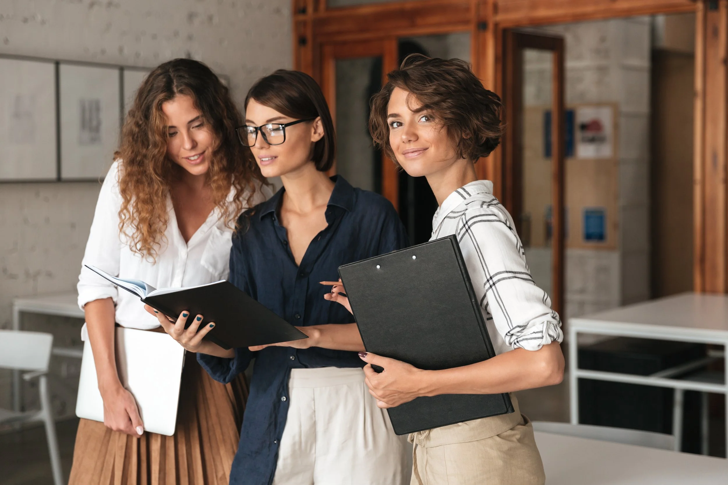 Three women standing together in an office, examining a black folder and holding notebooks.