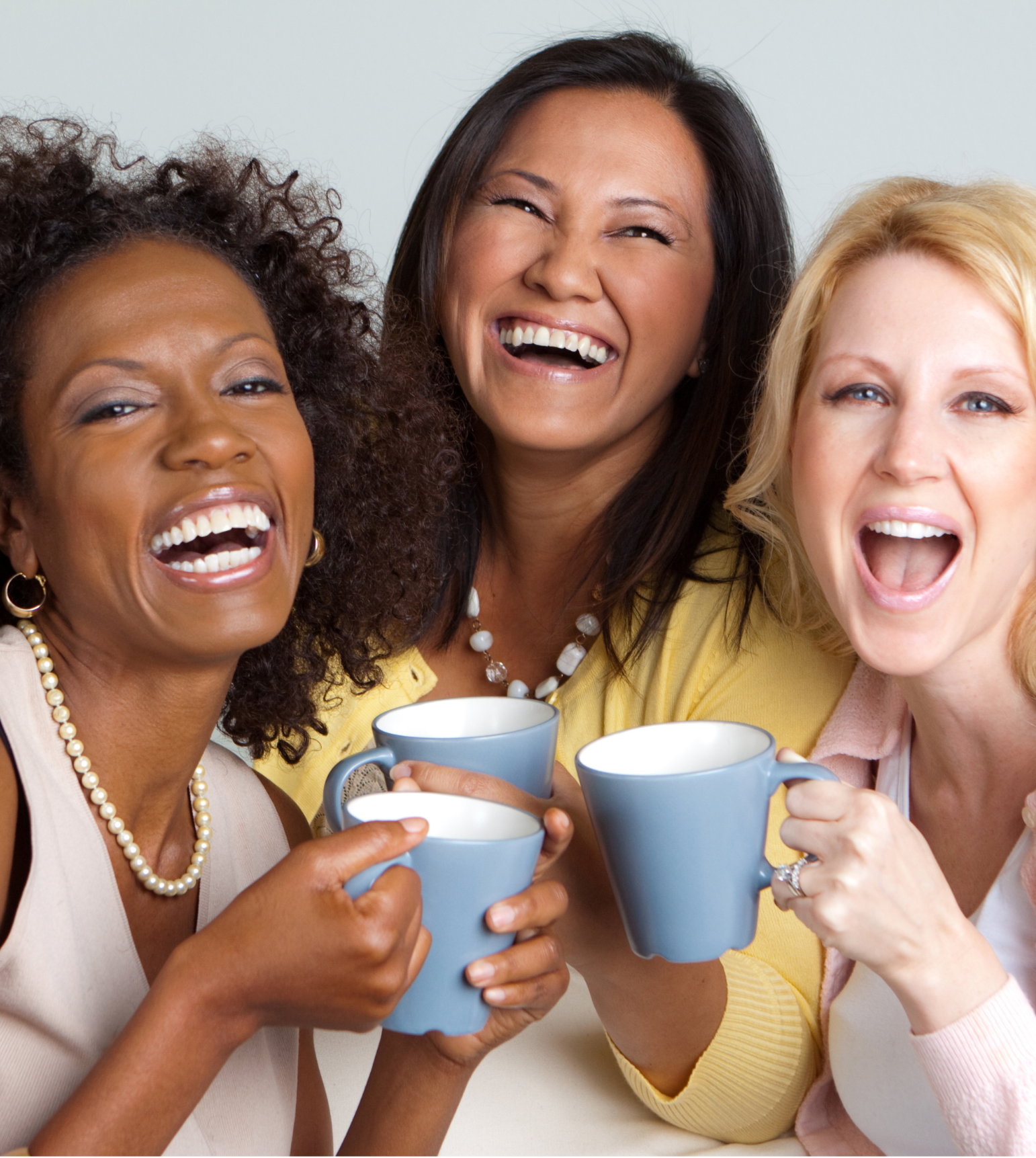 Three women laughing and holding blue coffee mugs together.