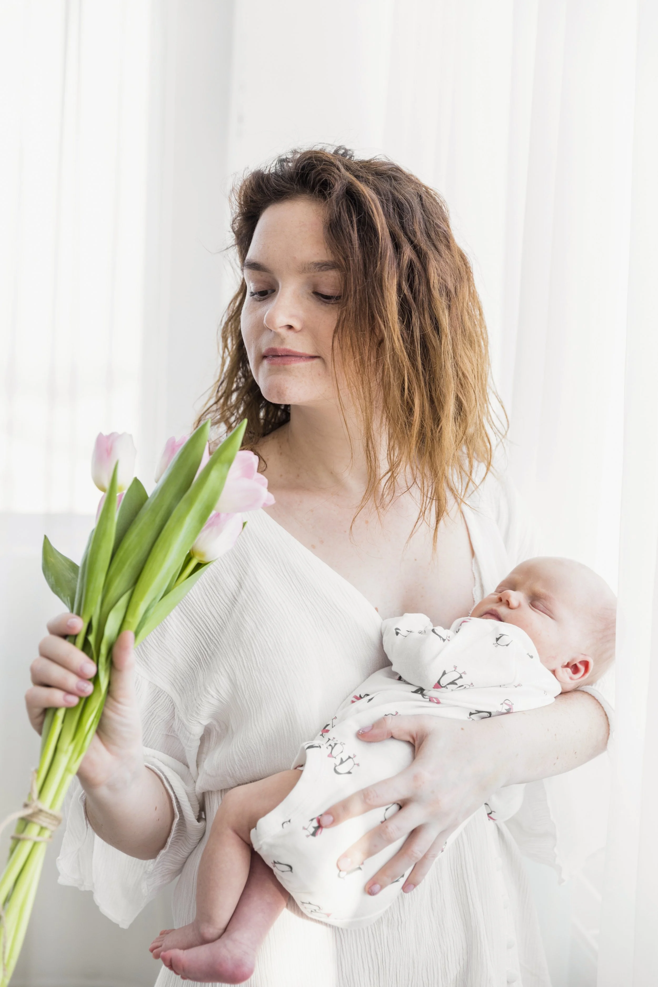 A woman with curly red hair holding a sleeping baby in her arm while looking at a bouquet of pink and white tulips.