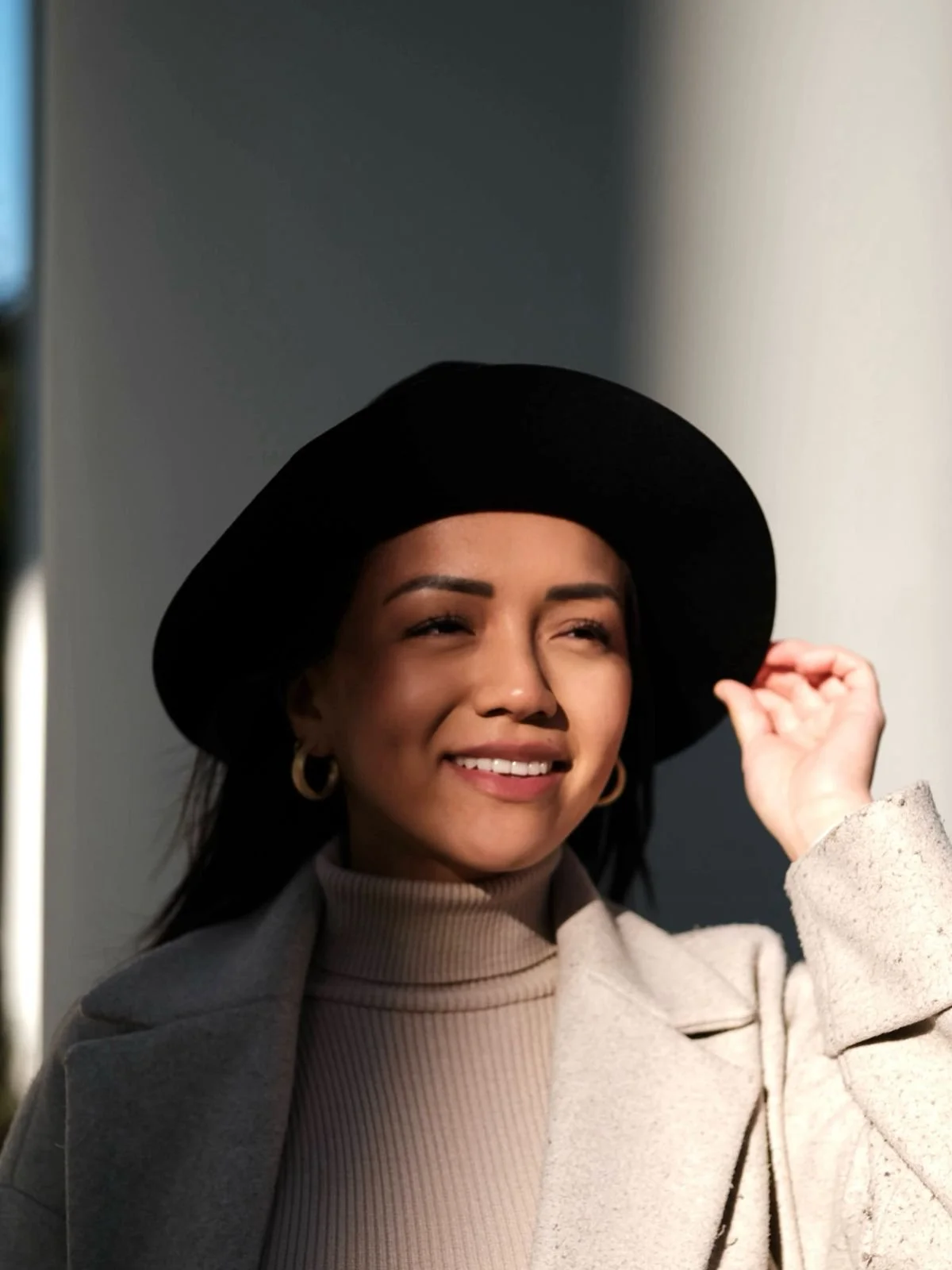 A woman with dark hair, wearing a beige turtleneck, hoop earrings, and a black hat, smiling and adjusting her hat in sunlight.