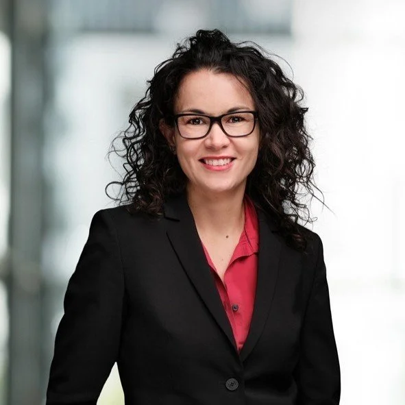 A woman with curly dark hair and glasses wearing a black blazer and red blouse, smiling in a professional indoor setting.