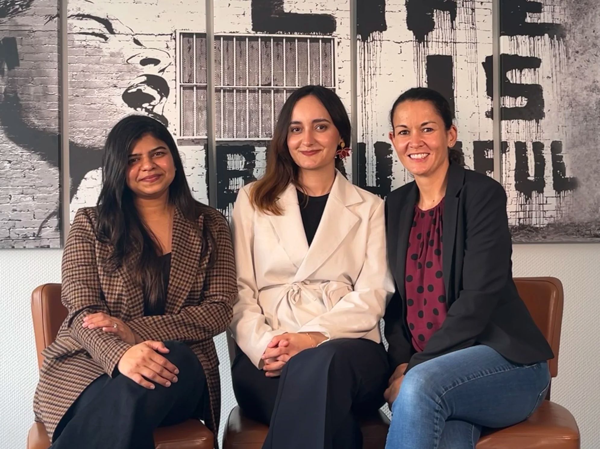 Three women sitting on a brown leather bench in front of a wall with a black and white mural of a face and the word "LOVE" painted in black.