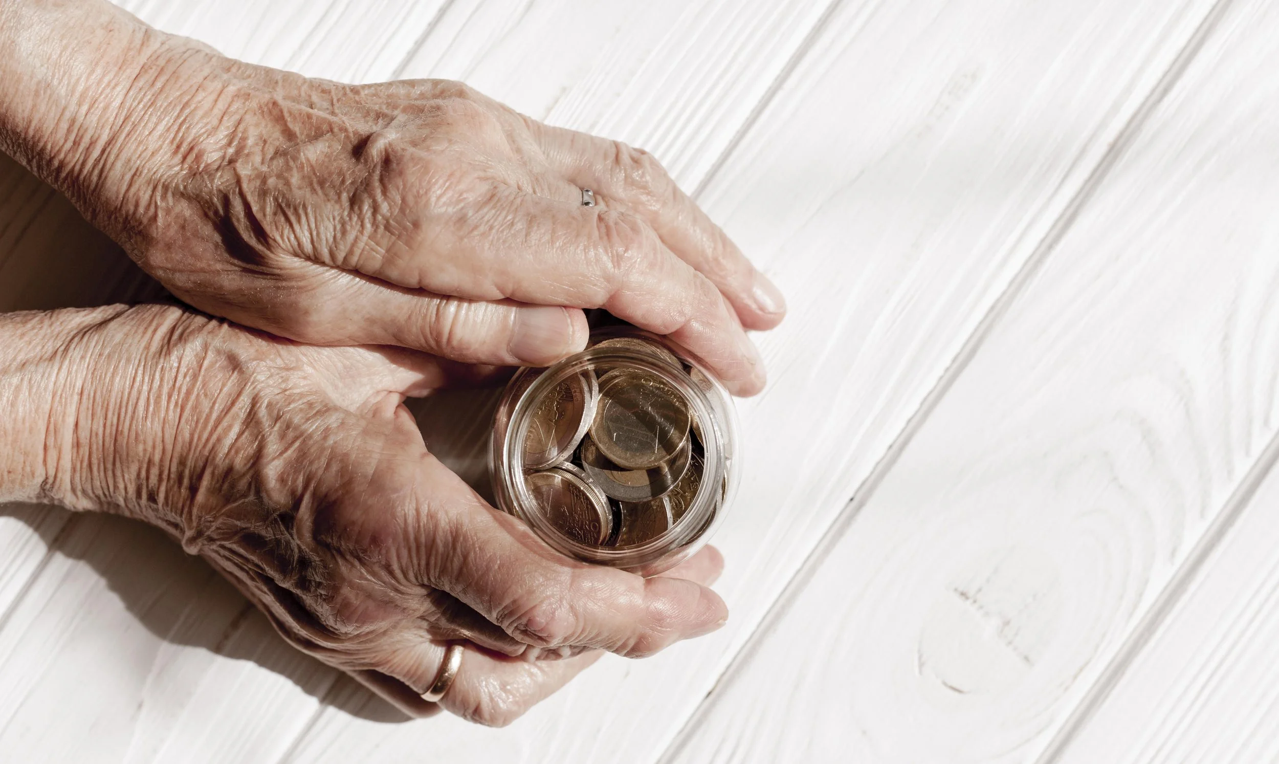 Elderly hands holding a jar of coins on a light wooden surface.