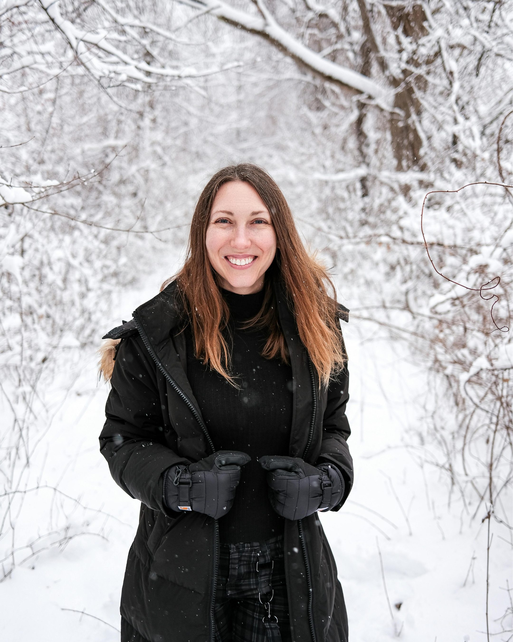 A woman smiling in a snowy outdoor setting, wearing a black jacket and gloves.