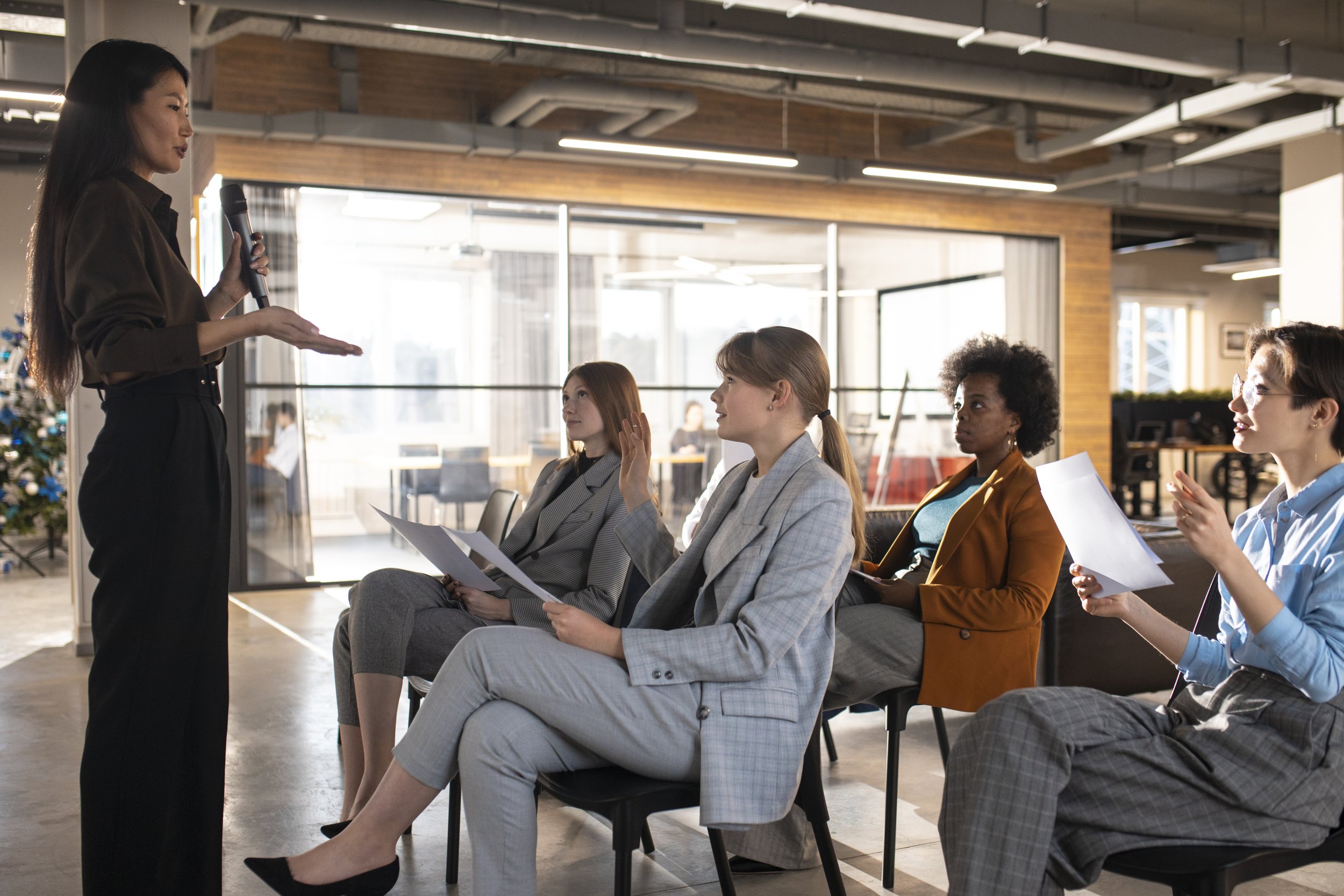 A woman standing and speaking into a microphone while a group of four women sitting and listening, some with papers, in a modern office conference room with large windows and a Christmas tree in the background.