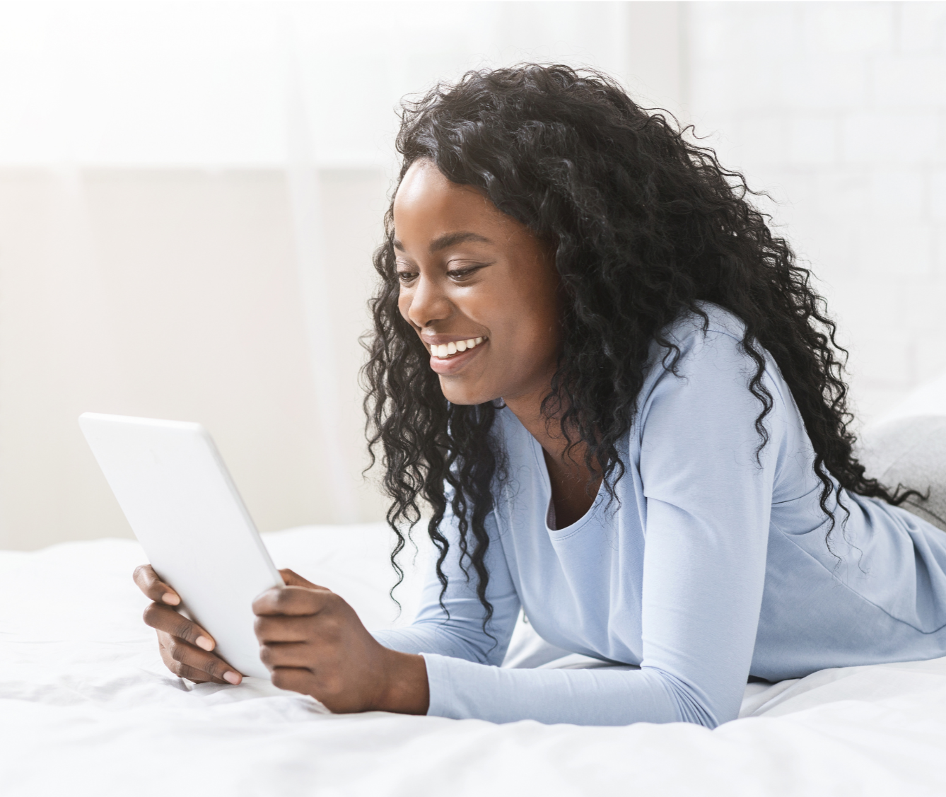 A woman with curly black hair smiling while lying on her stomach on a bed, holding a tablet in her hands.
