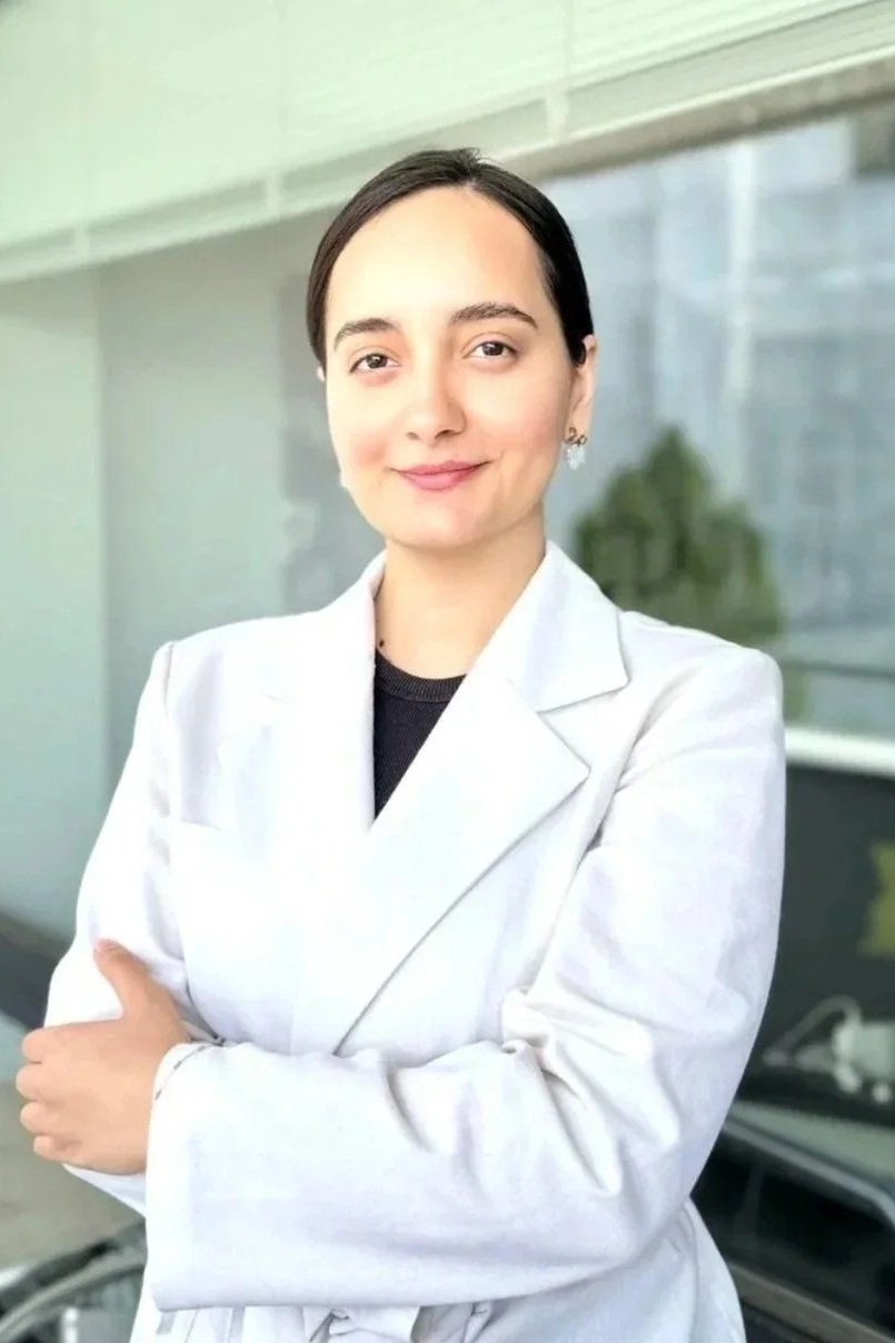 A woman in a white lab coat standing outside of a building, smiling with arms crossed.