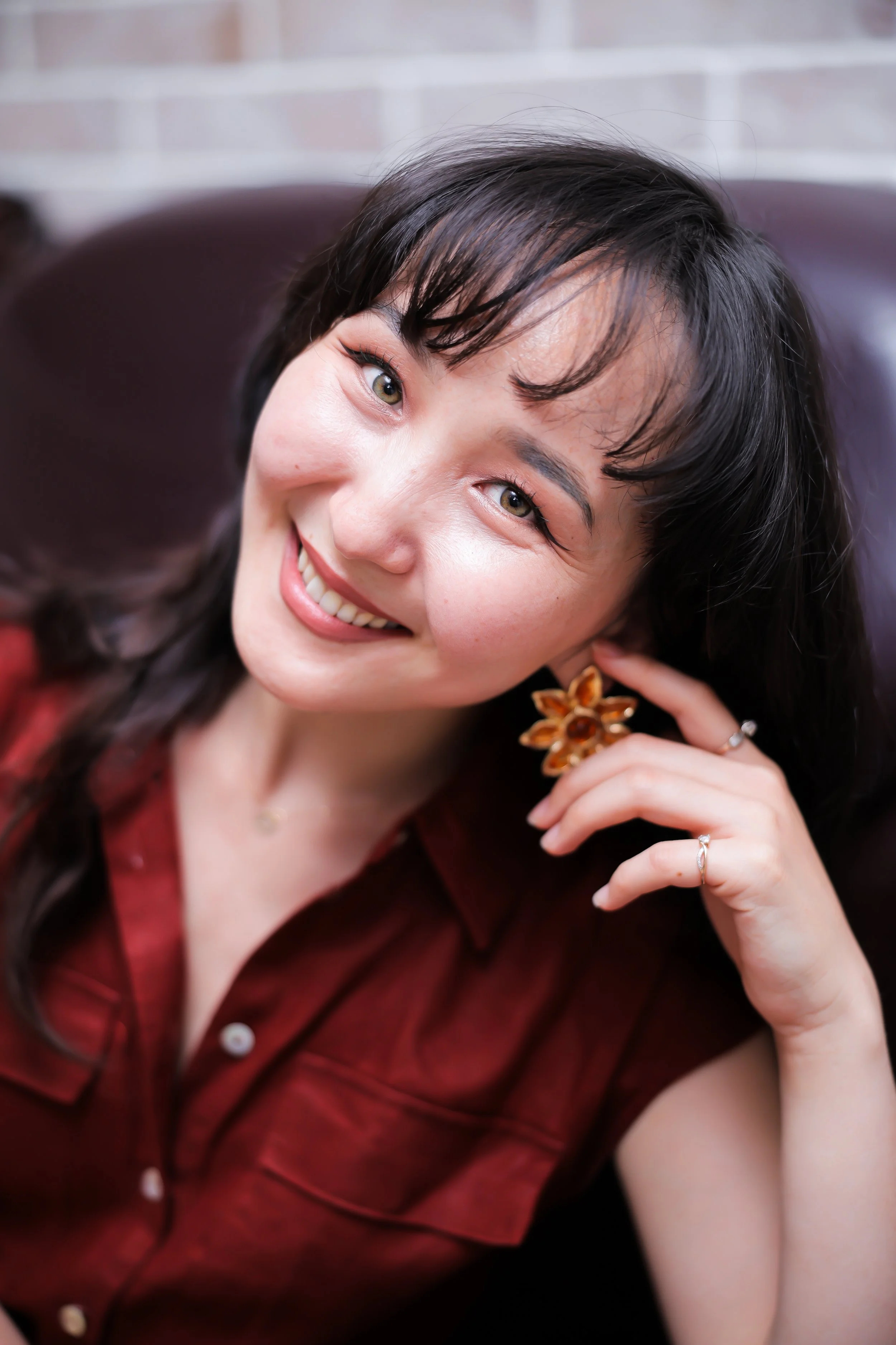 A smiling woman with dark hair and light skin, wearing a maroon shirt and sunflower-shaped earrings, holding her hand to her ear.