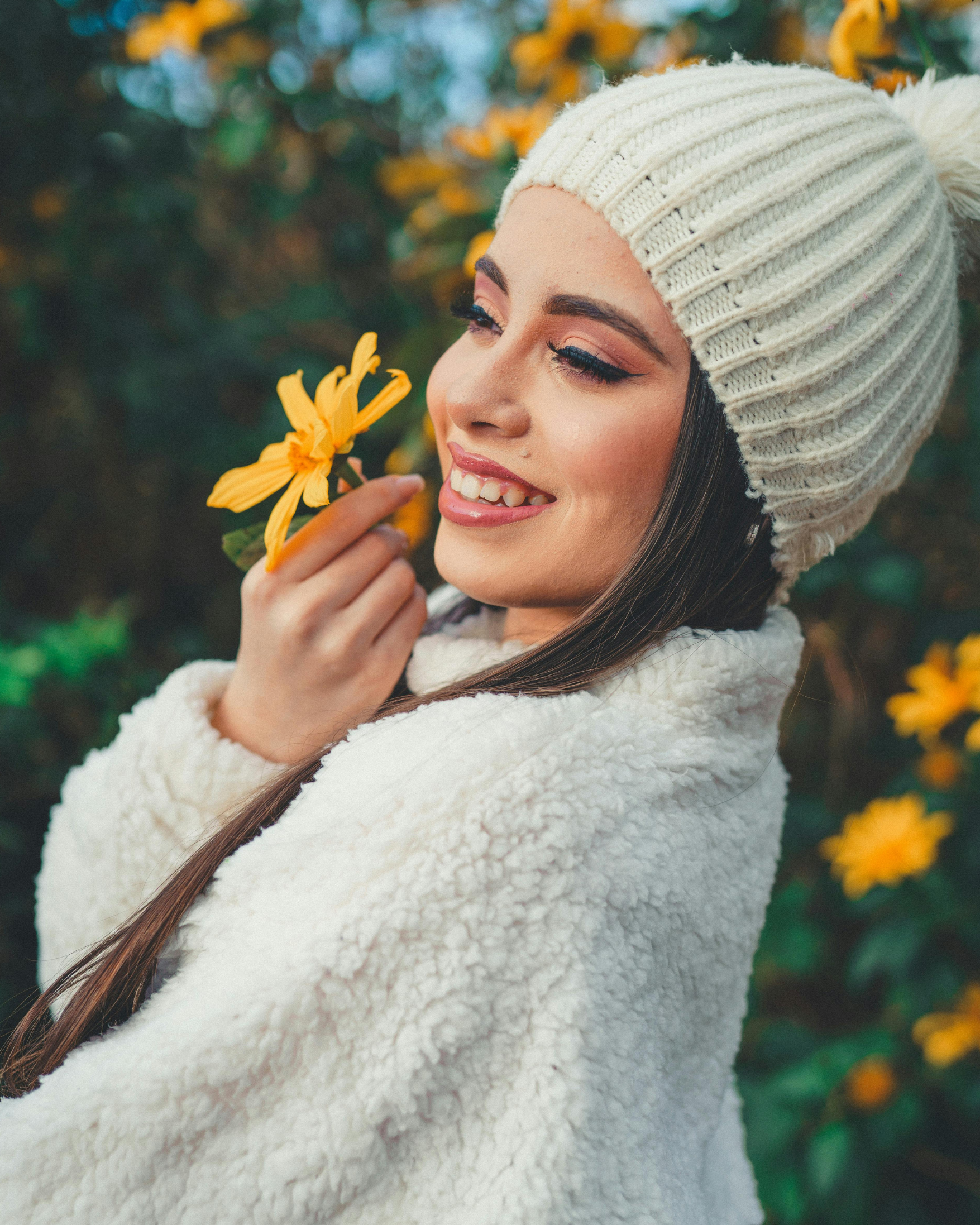 A woman smiling and holding a yellow flower close to her face outdoors, wearing a cream-colored knit hat and a white plush jacket.