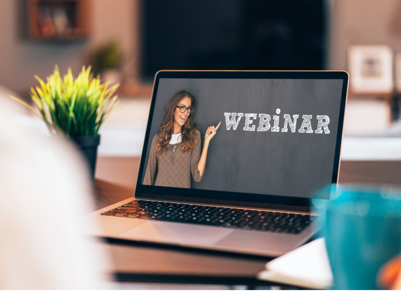 Laptop screen displaying a woman pointing at the word 'WEBINAR' on a chalkboard background, with a blurred background of a cozy home or office space.