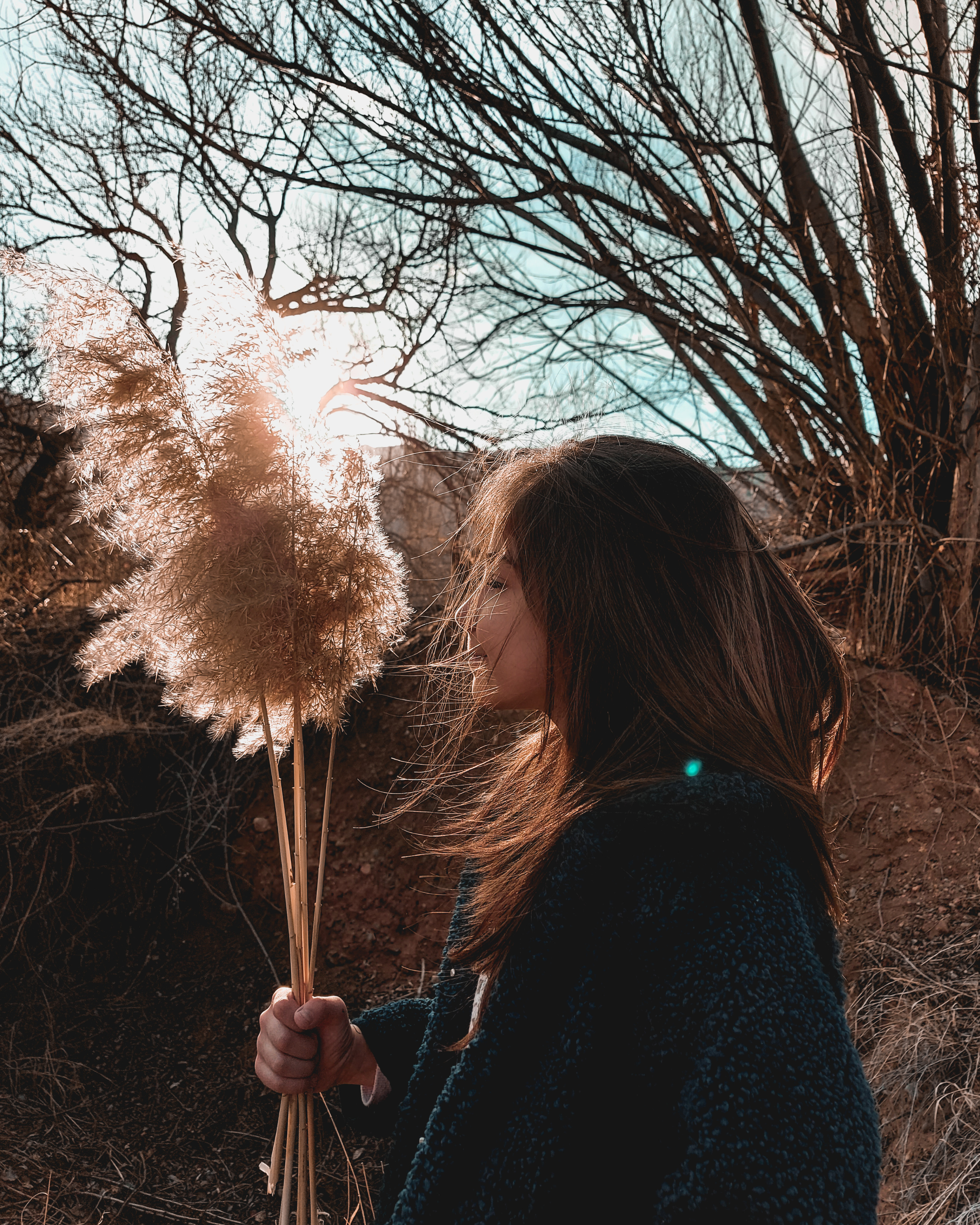 A woman with long brown hair holding a bundle of dried pampas grass in an outdoor setting with leafless trees and clear sky in the background.