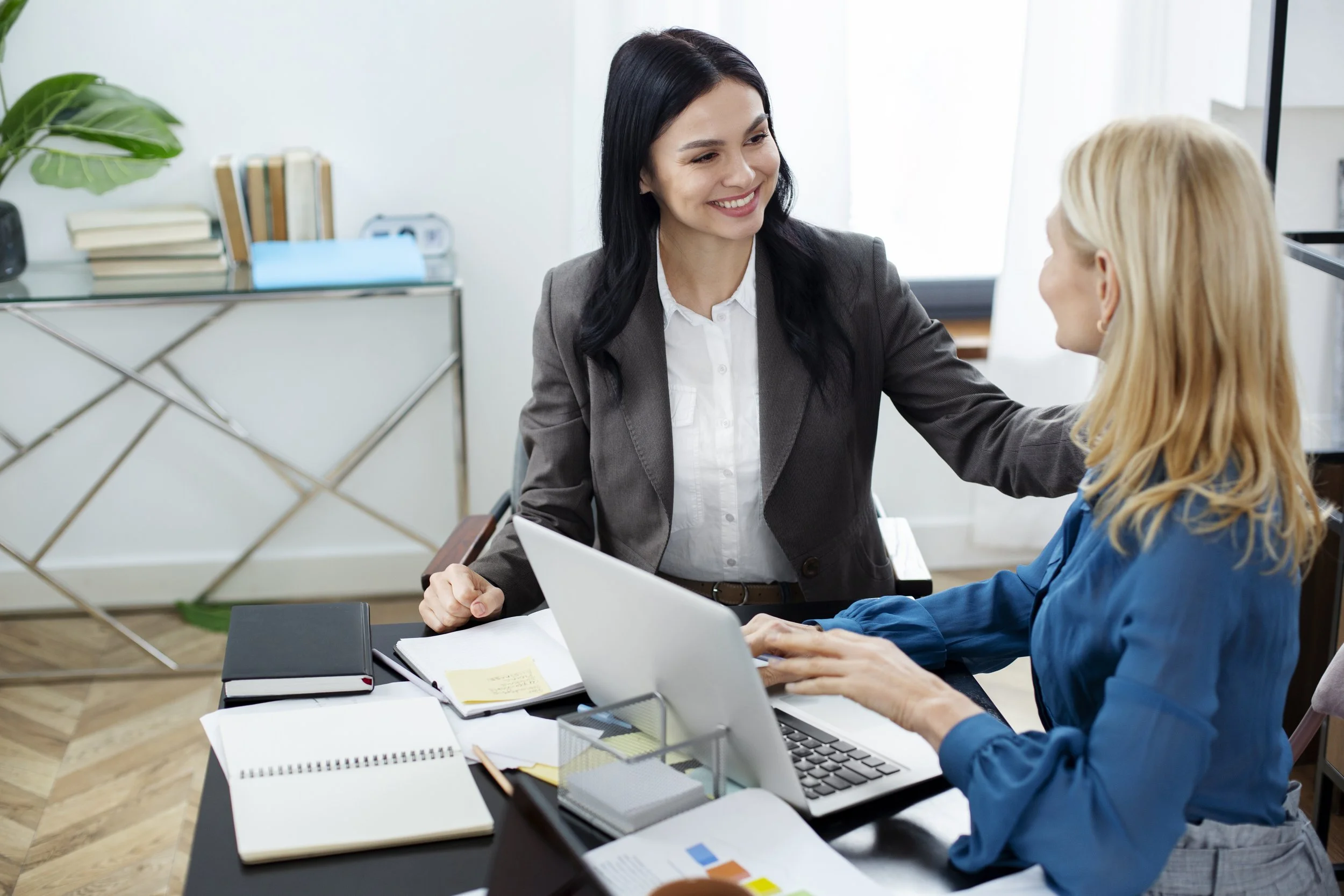 Two women in a professional office setting, one sitting at a desk and greeting the other with a smile, as they shake hands.