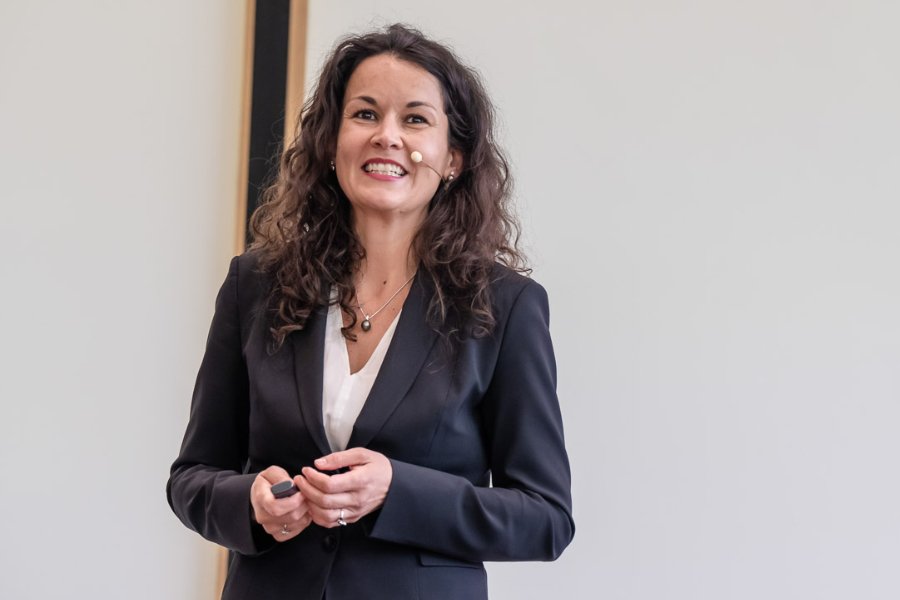 A woman with curly hair smiling, wearing a black blazer, white blouse, and necklace, giving a presentation or speech with a microphone headset.