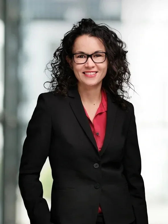 A woman with curly dark hair, glasses, and a friendly smile, dressed in a black blazer and a red shirt, standing in a modern office building with large windows in the background.