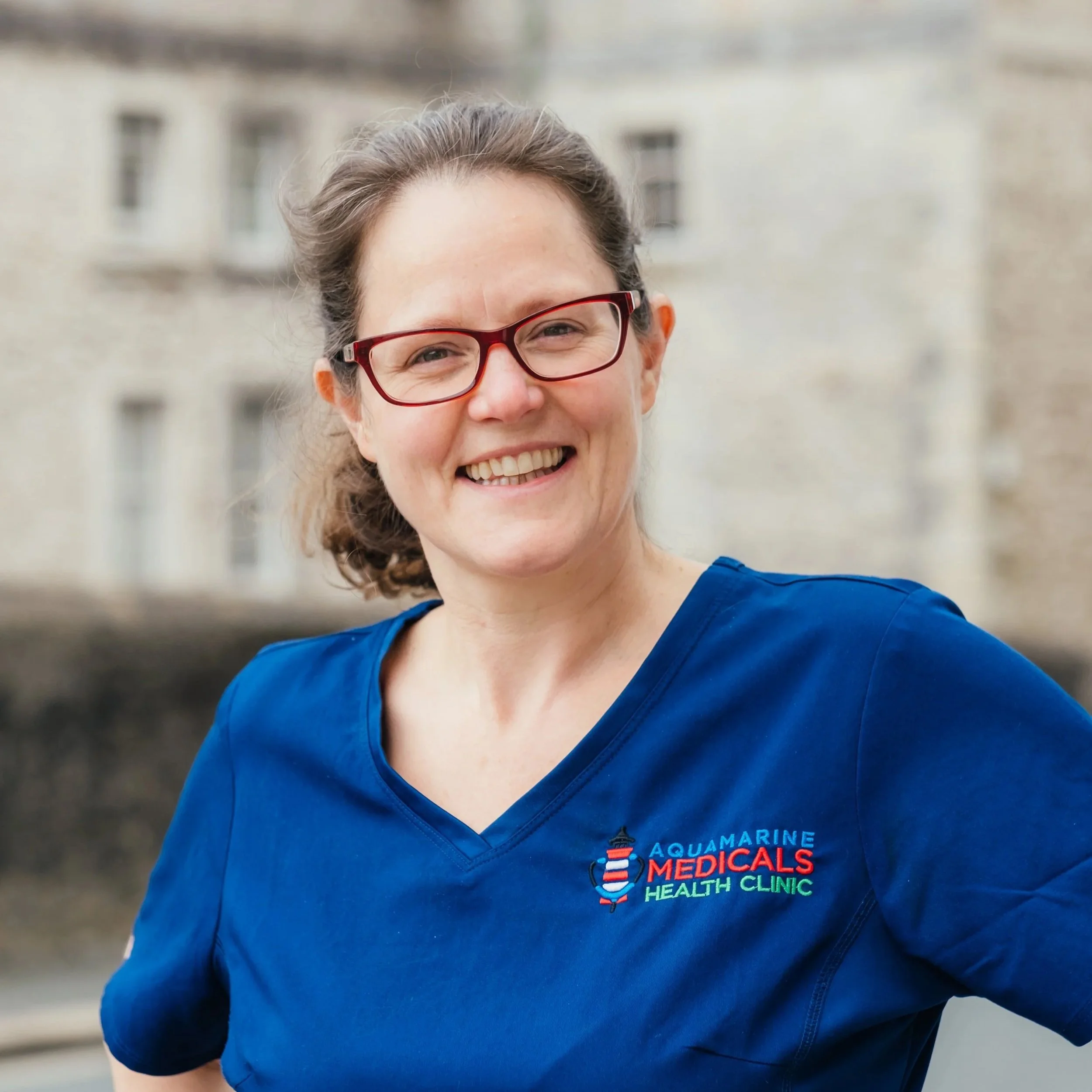 A smiling woman wearing red glasses and a blue medical uniform with the logo of Aquamarine Medicals Health Clinic, standing outdoors with a blurred building in the background.