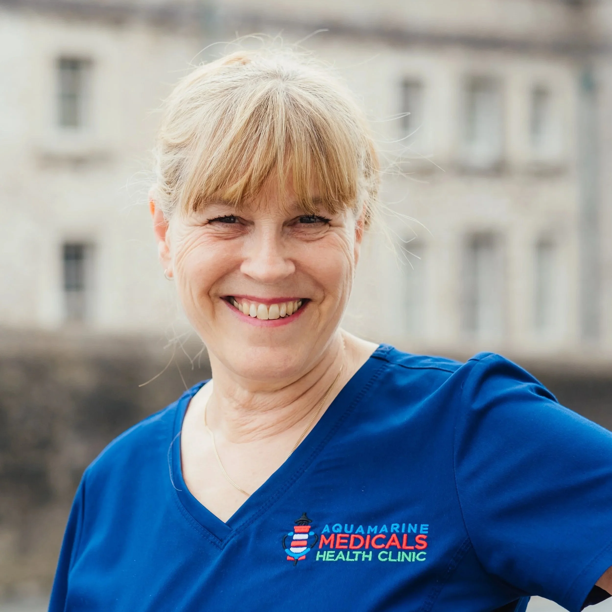 A smiling woman with blonde hair in a ponytail, wearing a blue medical scrub top with the logo for Aquamarine Medicals Health Clinic. She is standing outdoors with a blurred building in the background.
