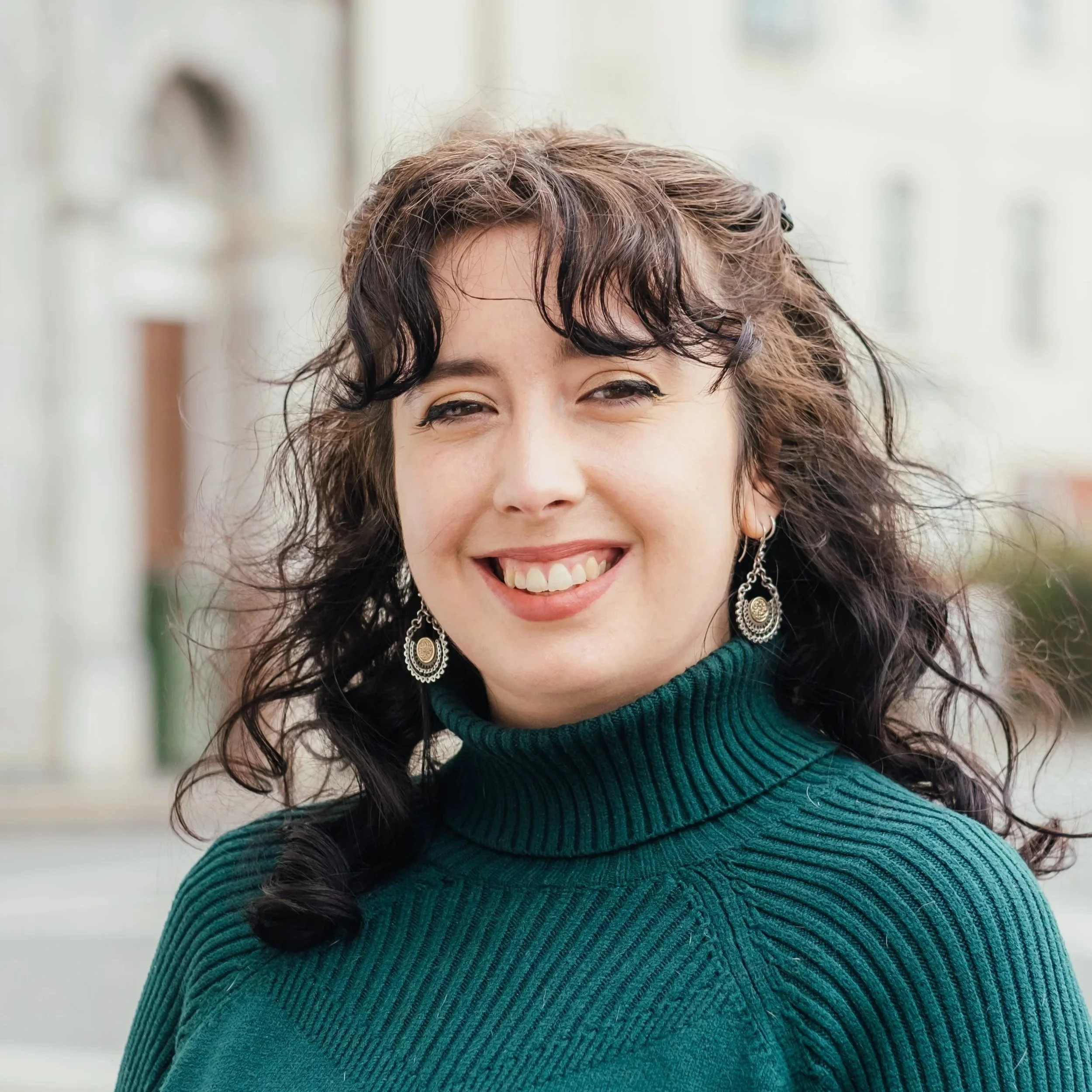 A woman with dark and curly hair, smiling, wearing a green turtleneck sweater and dangling earrings, outdoors with blurred buildings in the background.