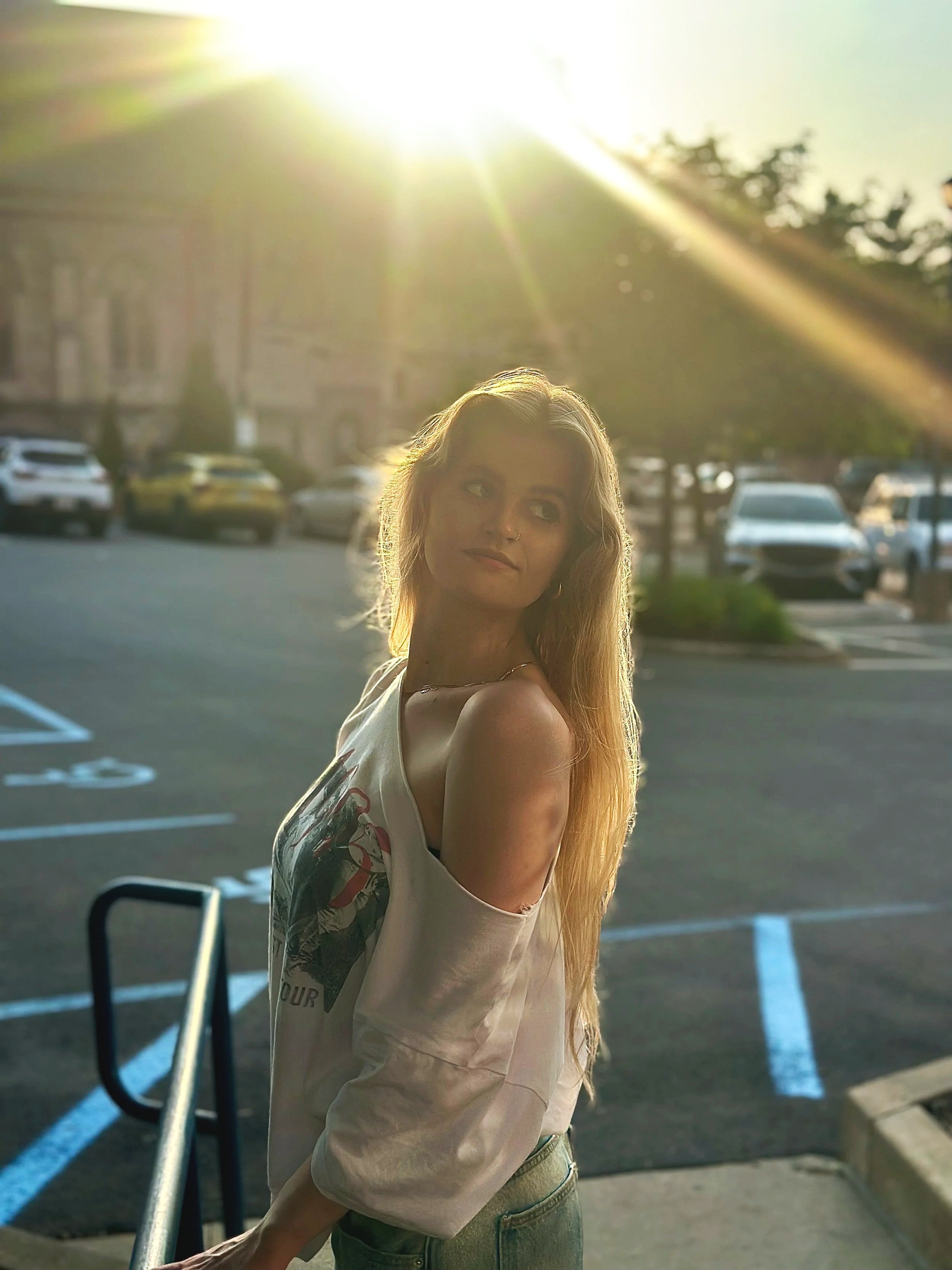 A young woman with long blonde hair standing in a parking lot during sunset, wearing a white off-shoulder shirt with a graphic design, looking to the side with a thoughtful expression.