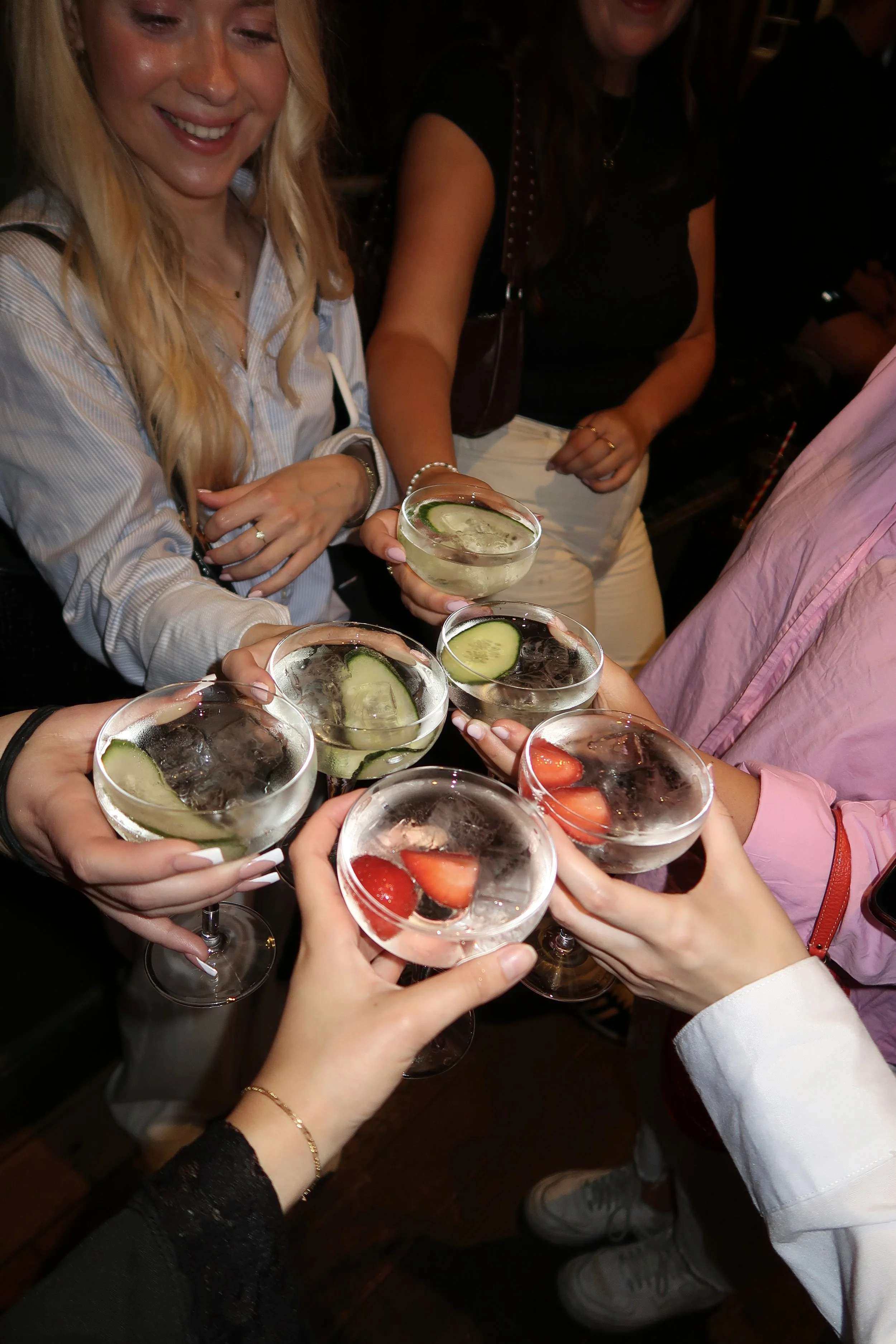 Group of people holding cocktails with fruit garnishes in a celebratory toast in a dark moody bar.