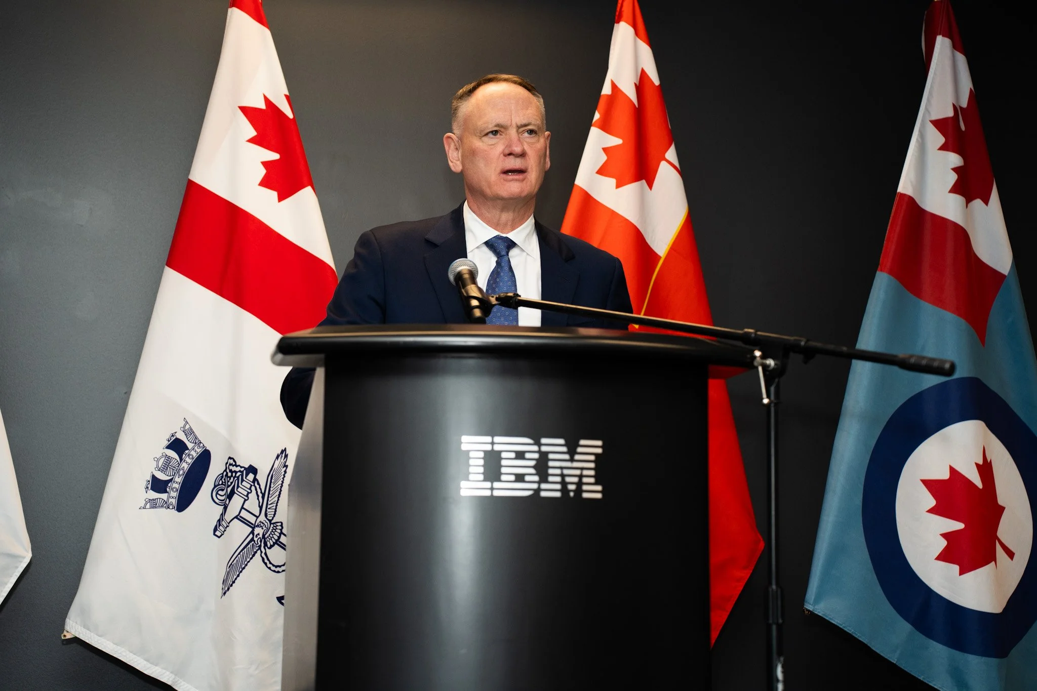 A man in a dark suit and blue tie speaking at a podium with the IBM logo, with Canadian flags behind him.