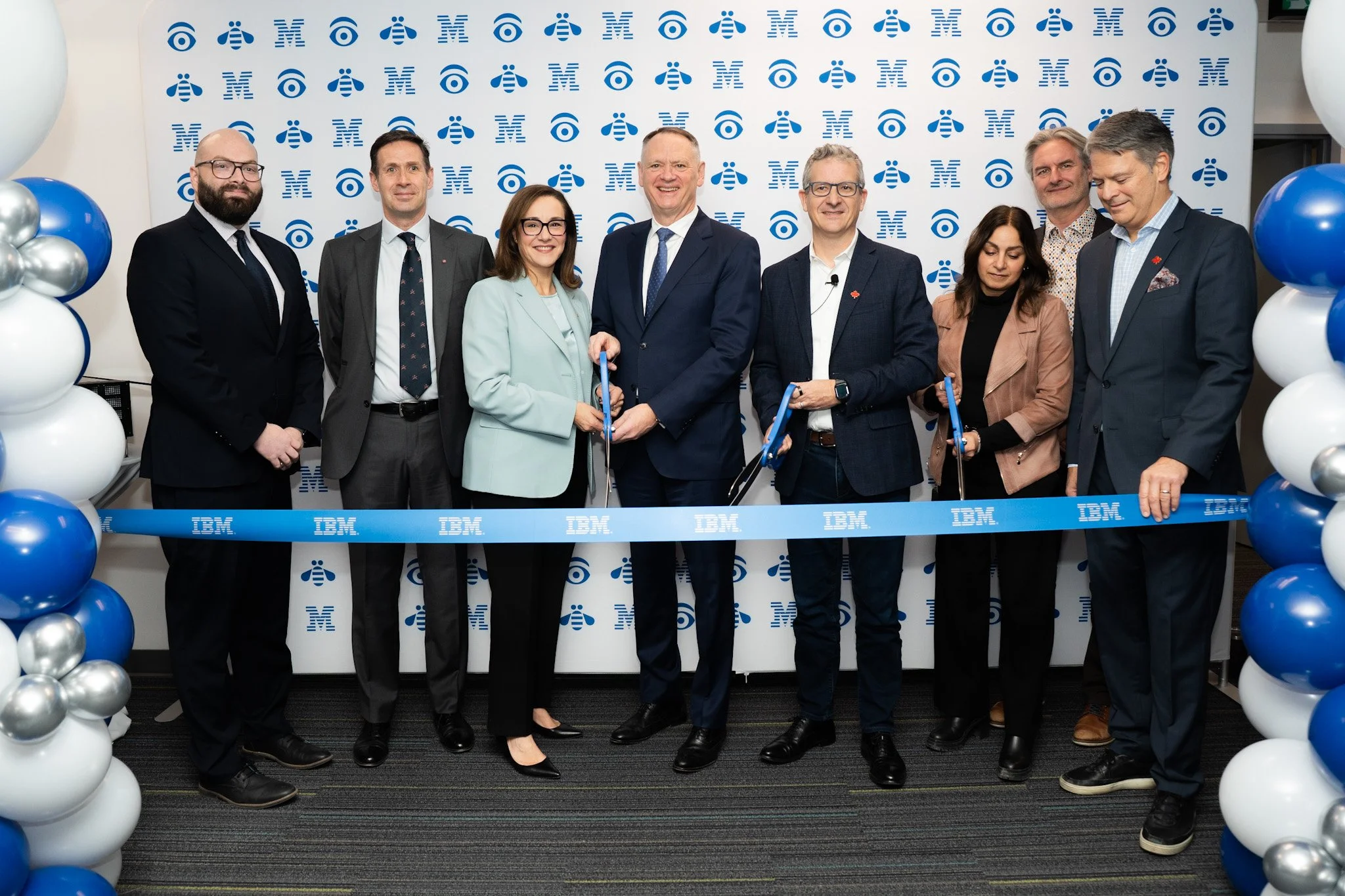 Group of eight professionals at a ribbon-cutting event at IBM, with a backward IBM logo background, holding scissors and flags.
