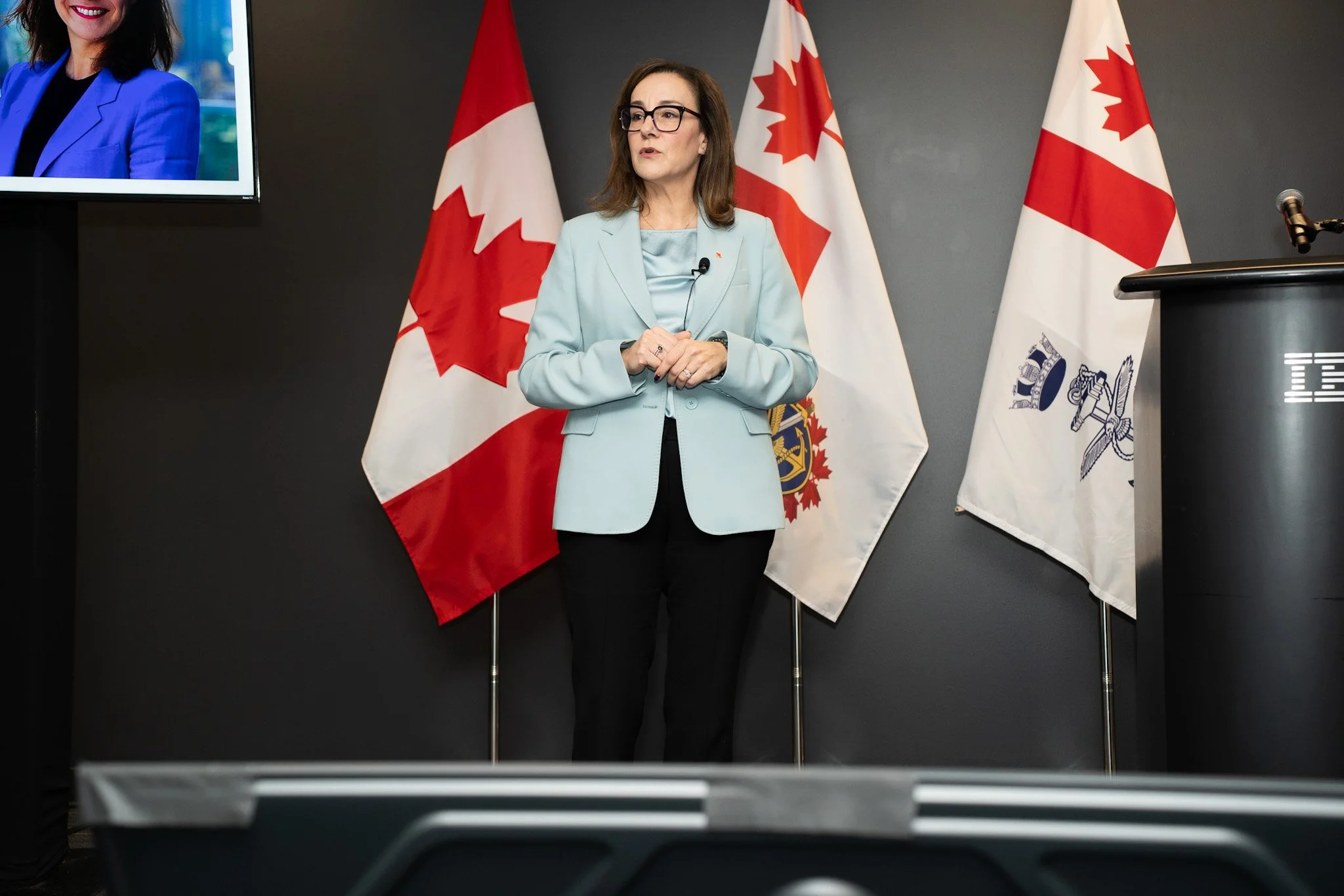 A woman standing at a podium with multiple Canadian flags behind her, speaking at an event.