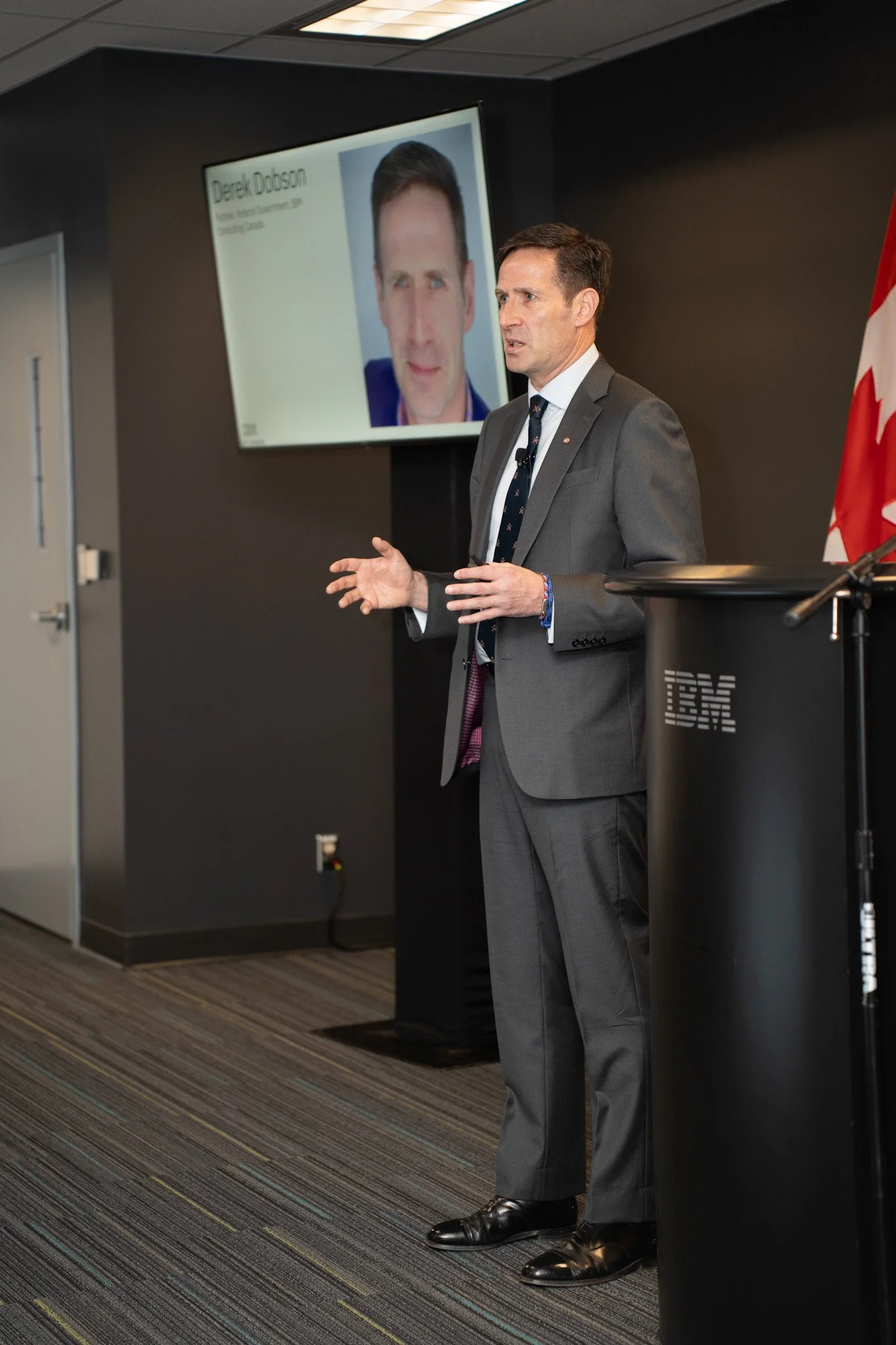 A man in a gray suit giving a presentation at a podium with an IBM logo. There is a large screen behind him displaying his image and the name Derek Dobson.