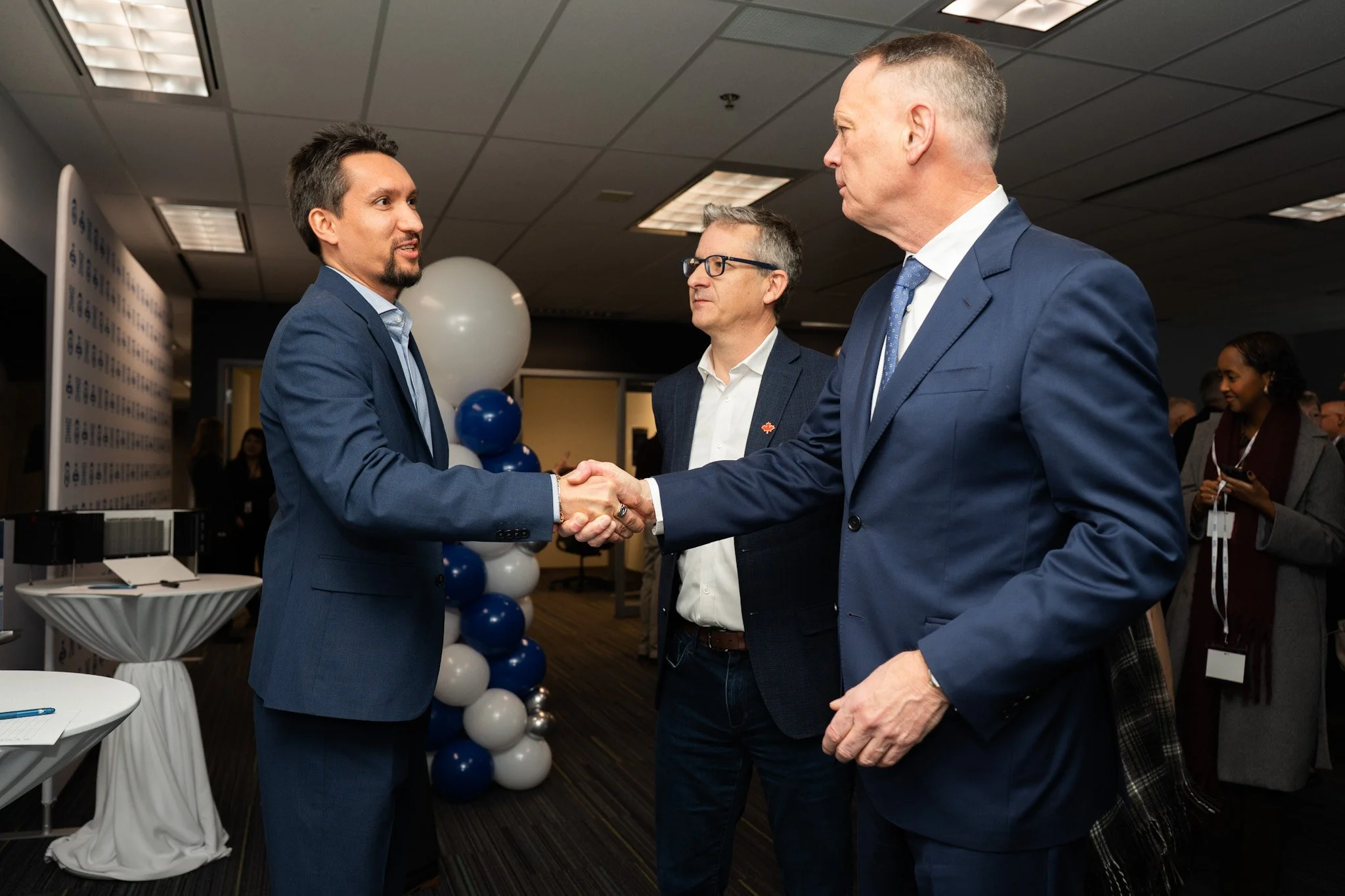 Two men in suits shaking hands during a formal event, with a third man observing in the background. Balloons and event decorations are visible.