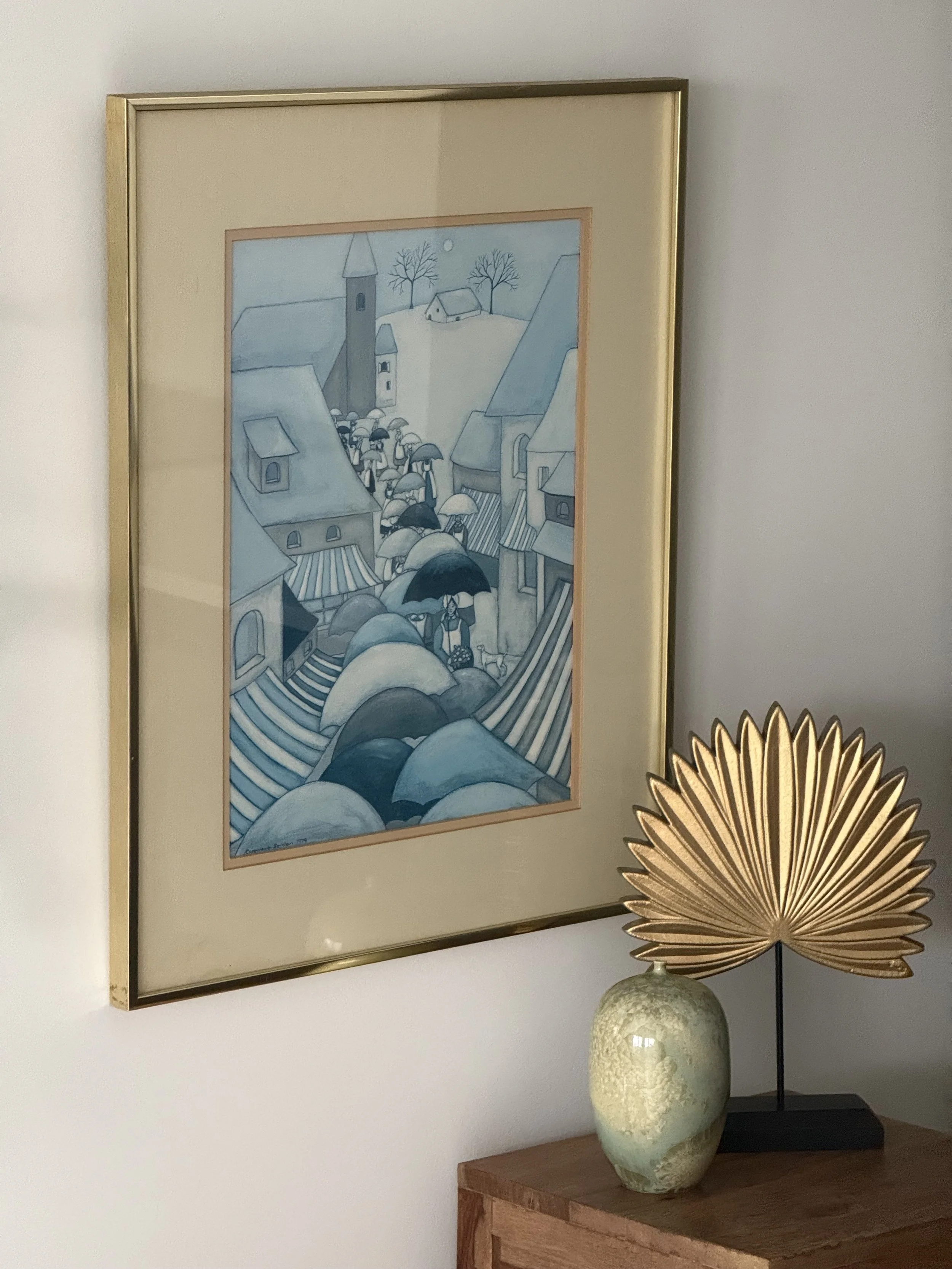 Framed vintage naïve folk art drawing depicting a blue-toned village procession with umbrellas, signed 1979, matted and set in a slim gold-tone frame, shown styled on a wall above a wooden surface