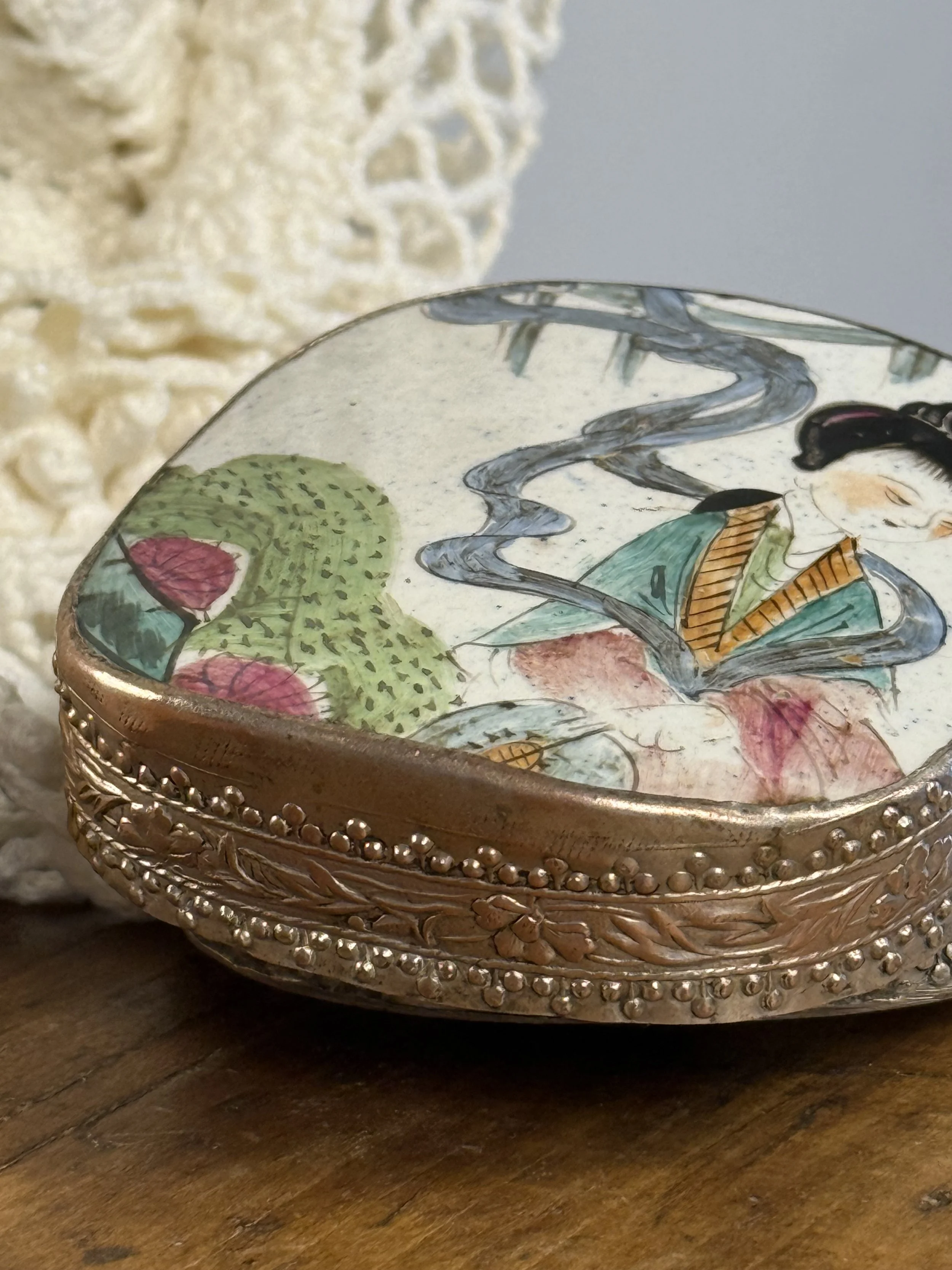 Side view of vintage porcelain and silver-toned trinket box highlighting repoussé floral banding, raised dot detailing, and softly worn patina along the metal edge