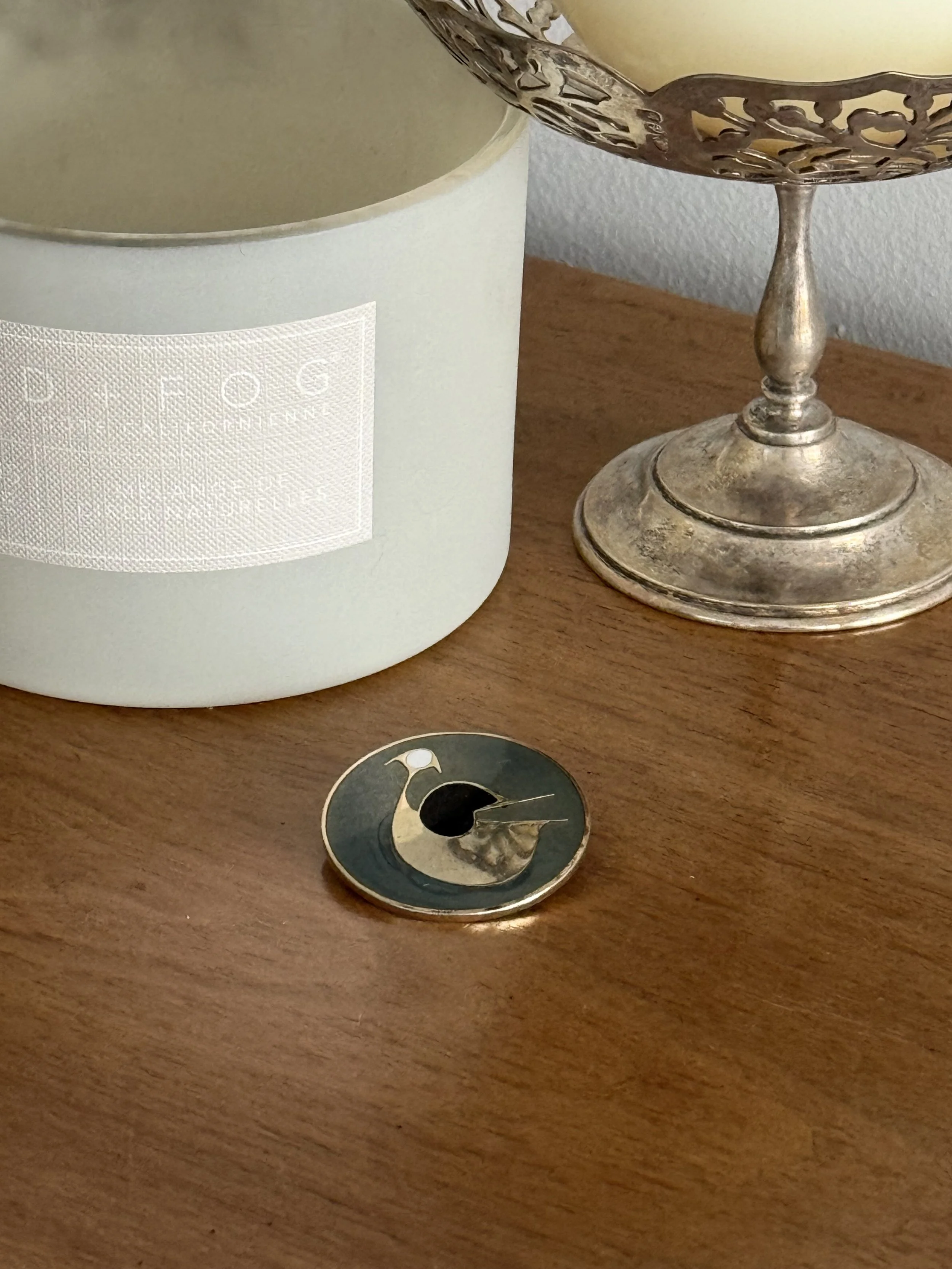 Mid-century modern brass bird brooch styled on wooden surface beside silver compote and candle, sculptural enamel bird pin with organic silhouette