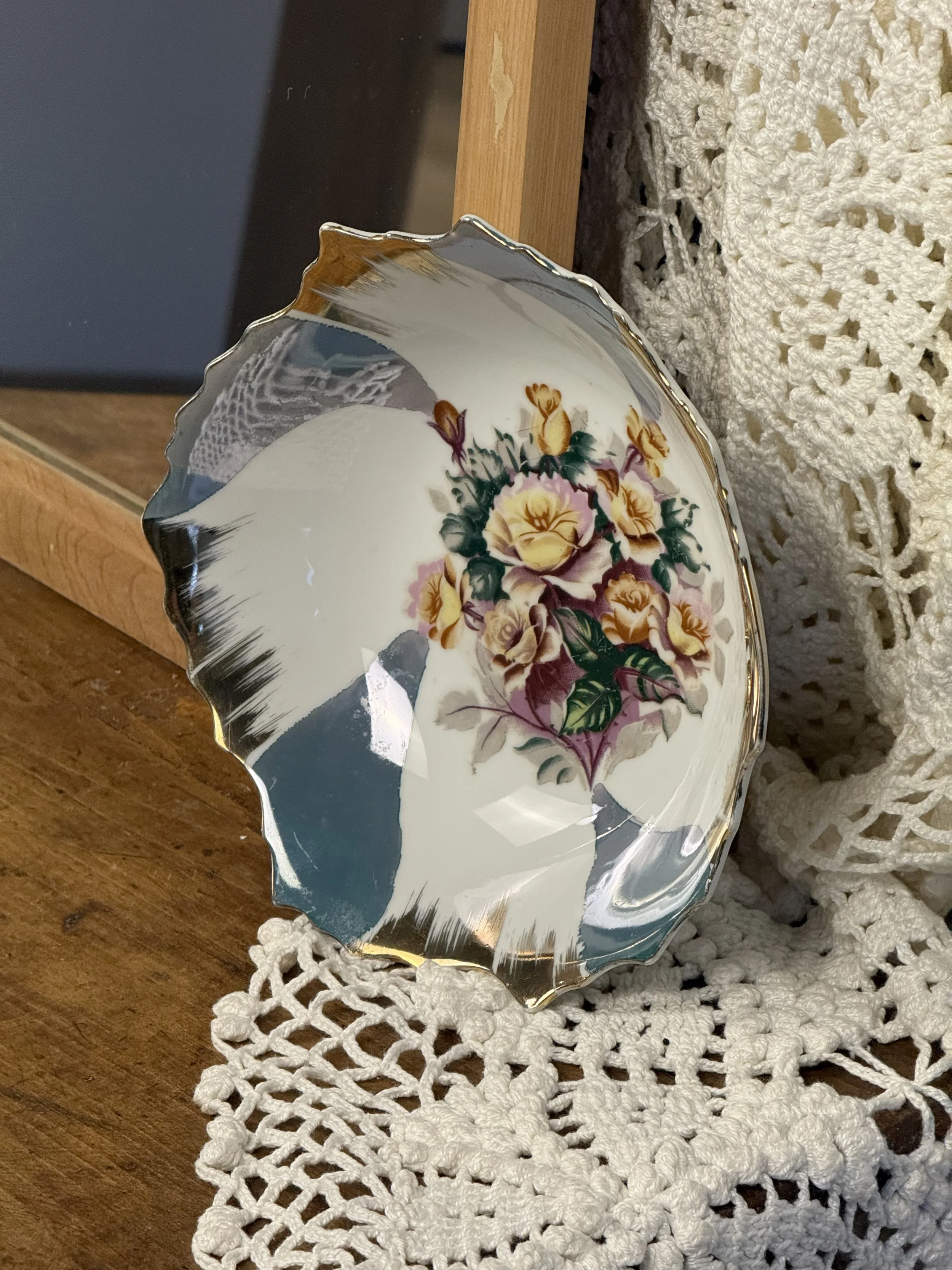 Three-quarter view of vintage porcelain plate leaning against wooden frame, featuring romantic rose bouquet motif, painterly glaze accents, and elegant scalloped silhouette