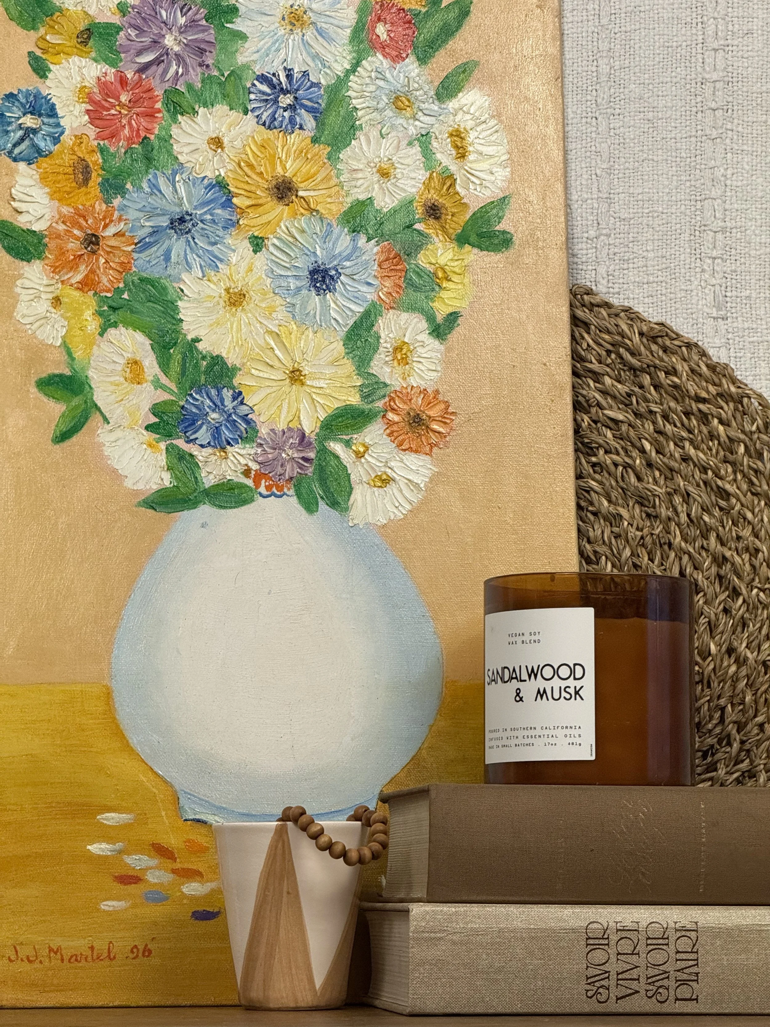 Styled scene with J.J. Martel’s 1996 folk art oil painting of impasto flowers in a pale vase on an ochre table, shown with books, beads, and a candle