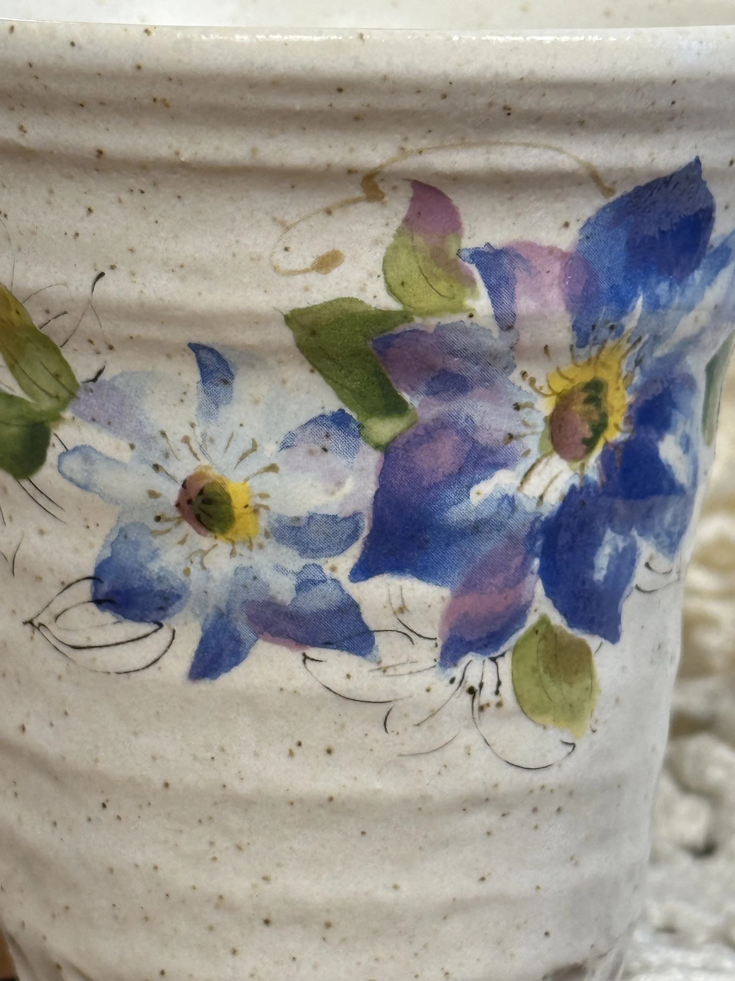 Close-up detail of hand-painted blue and violet flowers with green leaves on Japanese stoneware, visible brushwork and natural glaze speckling